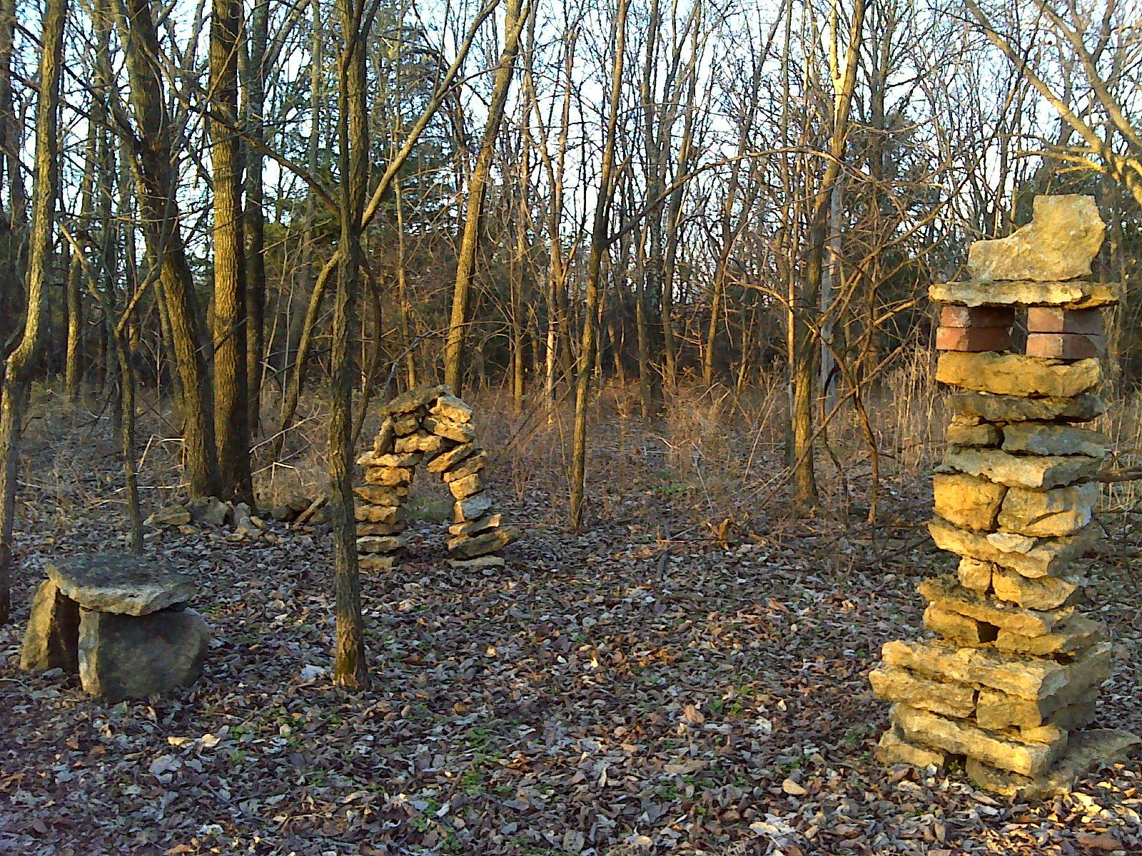 A woodland scene featuring several rock structures amidst bare trees. On the left, a low stone table is partially covered with dried leaves. In the center, an arched stone formation stands, and to the right is a tall stone pillar with bricks incorporated into its design. The ground is scattered with fallen leaves, indicating a late autumn or early winter setting. Lehigh Portland Trails mountain bike trail.
