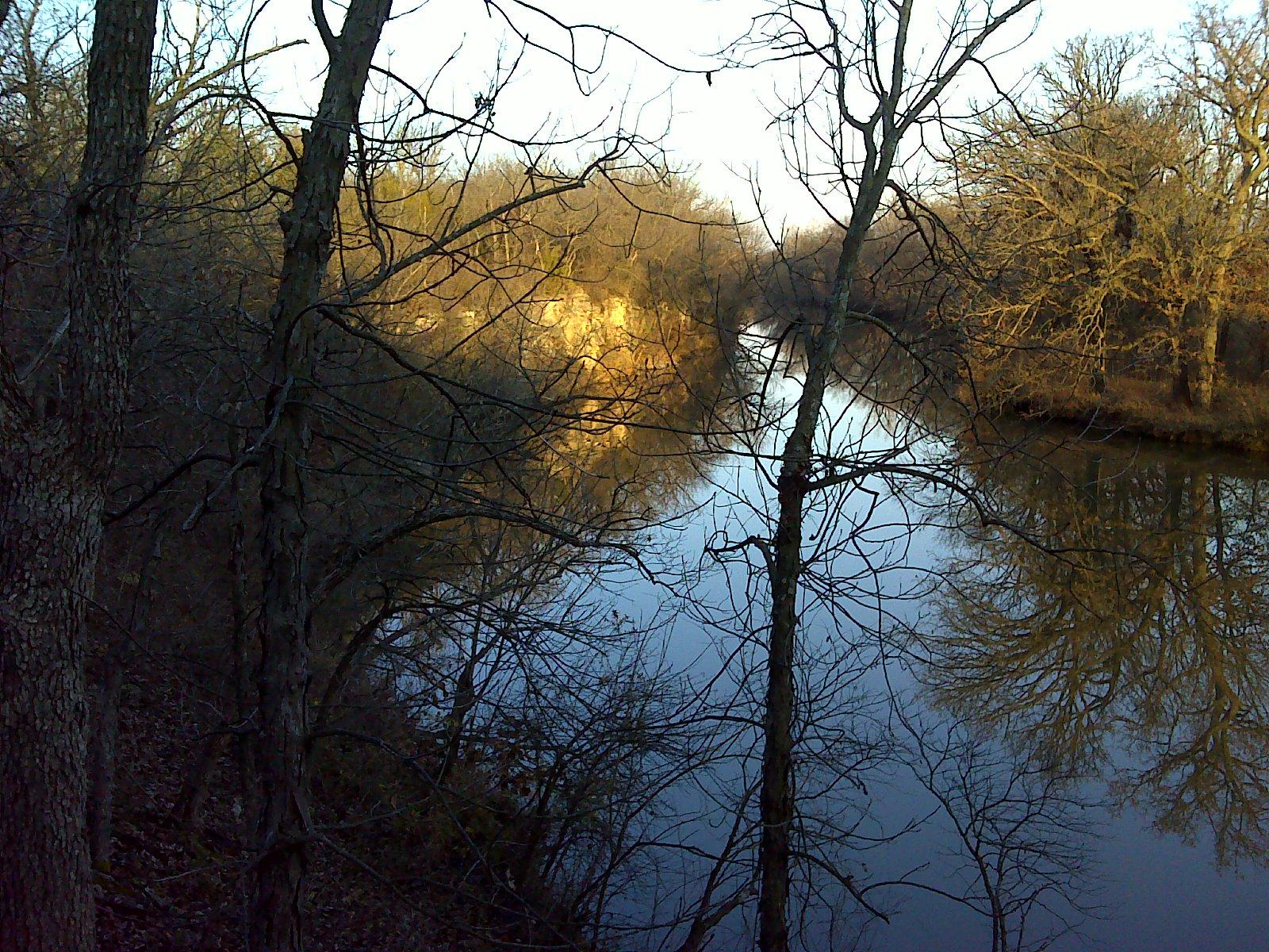 A tranquil river scene during early morning, framed by bare trees. The water reflects the calm surface and the surrounding landscape, which features patches of golden light on the riverbank and tree silhouettes. Lehigh Portland Trails mountain bike trail.
