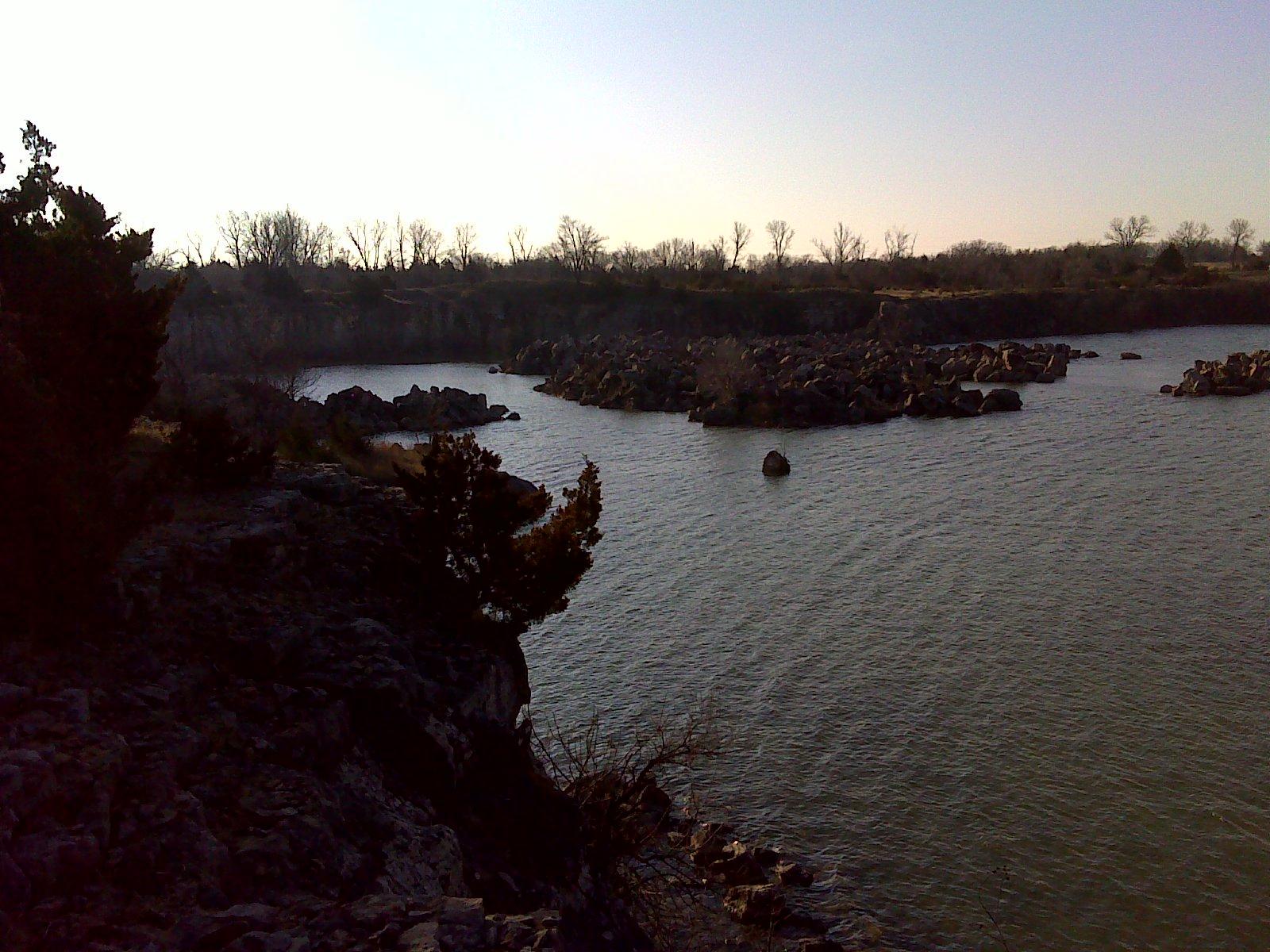 Rocky shoreline along a calm body of water, with trees in the background and boulders scattered across the water's surface. The scene is bathed in soft, natural light, suggesting early morning or late afternoon. Lehigh Portland Trails mountain bike trail.
