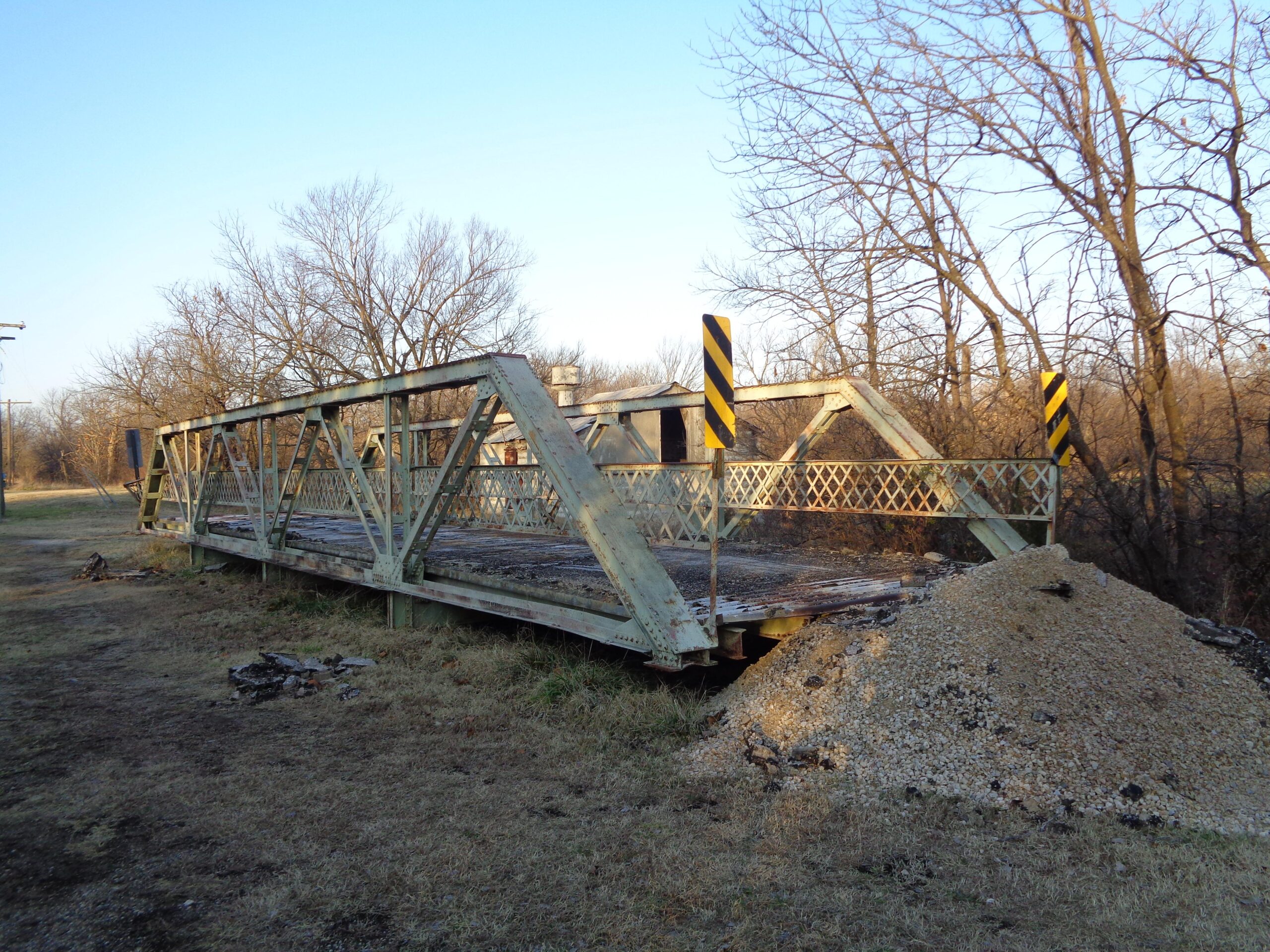 An old, rusted metal bridge sits unused, with a pile of gravel at its base. Surrounding the bridge are sparse trees and overgrown grass, under a clear blue sky. Black and yellow warning signs are positioned at either end of the bridge, indicating it may no longer be safe for passage. Lehigh Portland Trails mountain bike trail.