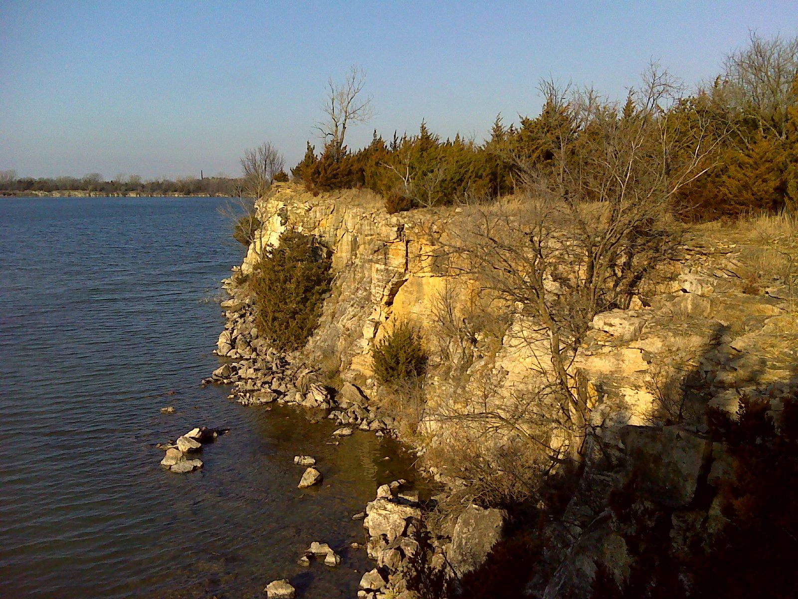Rocky shoreline with a cliff overlooking a calm body of water, surrounded by sparse trees and a clear blue sky. Small boulders are visible along the water's edge. Lehigh Portland Trails mountain bike trail.