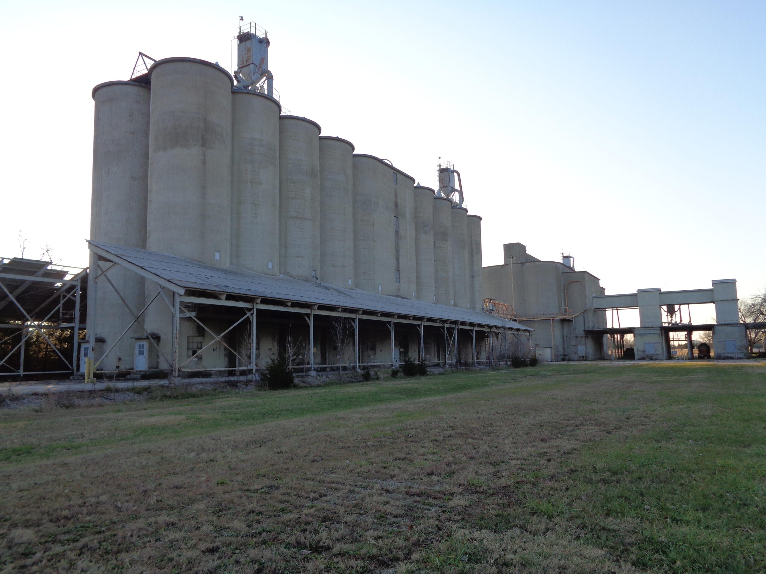 Alt text: A large industrial structure featuring several tall, cylindrical silos made of concrete, with an adjacent covered area supported by metal beams. The setting includes a grassy area in the foreground and a clear sky in the background. Lehigh Portland Trails mountain bike trail.