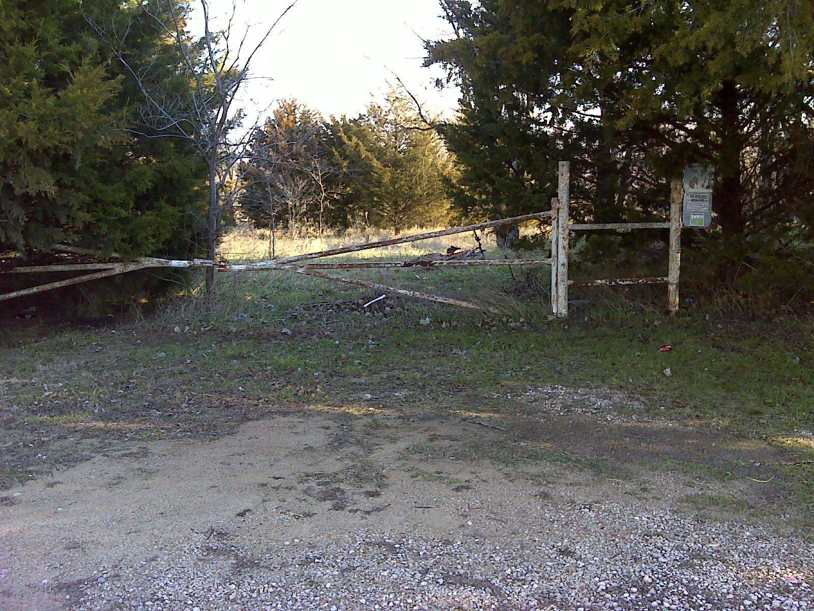 An old, weathered barbed wire gate stands partially open, surrounded by overgrown grass and trees. The gate is supported by a tree on one side and rusty metal posts. A sign is attached to one of the posts, indicating the area beyond the gate. The scene is set in a rural landscape with a dirt path leading toward the gate. Lehigh Portland Trails mountain bike trail.