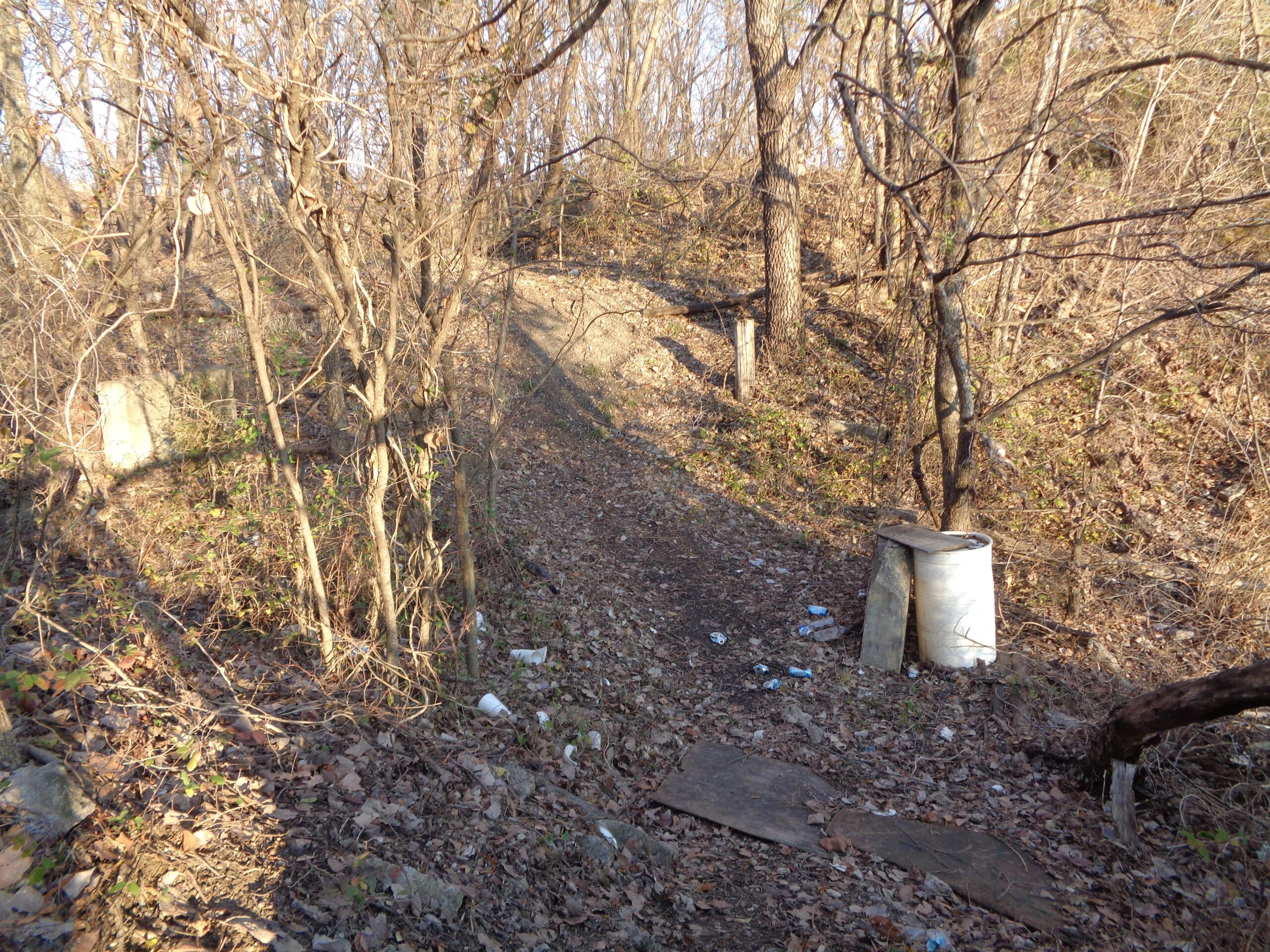A narrow, winding trail through a wooded area in late fall, with bare trees and fallen leaves scattered on the ground. To the right, there is a white barrel and a piece of wood, along with some litter visible along the path. The sunlight casts shadows, adding depth to the scene. Lehigh Portland Trails mountain bike trail.