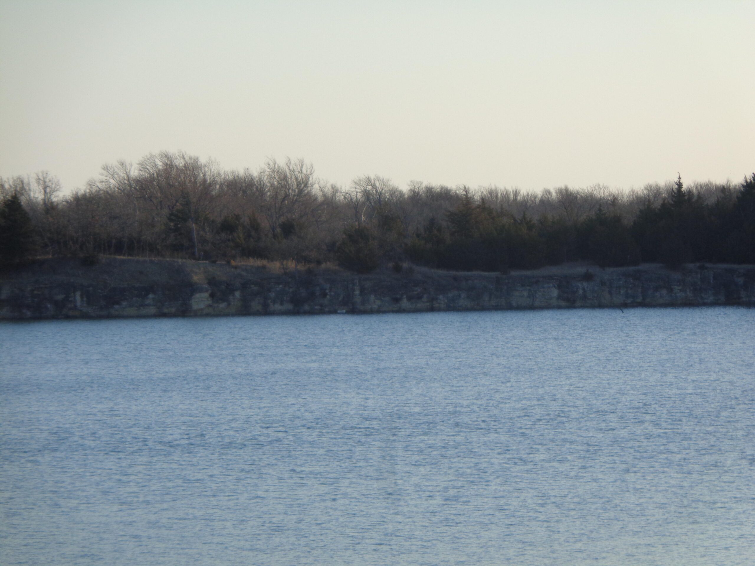 A tranquil view of a calm lake bordered by a rocky cliff and sparse tree line under a clear sky. The water reflects the surrounding landscape, creating a serene atmosphere. Lehigh Portland Trails mountain bike trail.