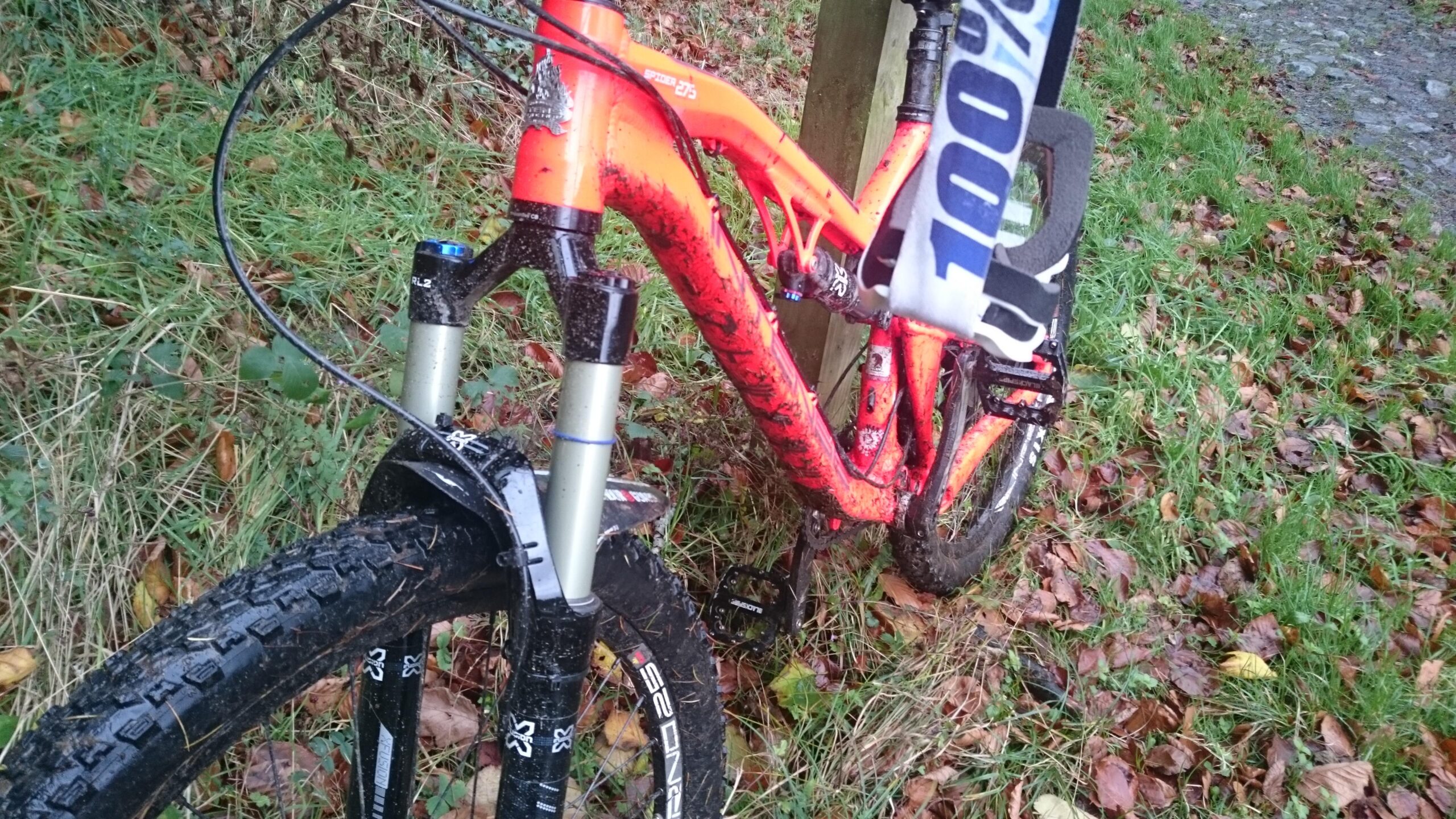 Intense Spider 275: A close-up of a mountain bike with an orange frame, partially covered in mud, parked near a grassy area with fallen leaves. The bike features thick tires and front suspension, indicating it is designed for off-road use. A section of a trail is visible in the background.