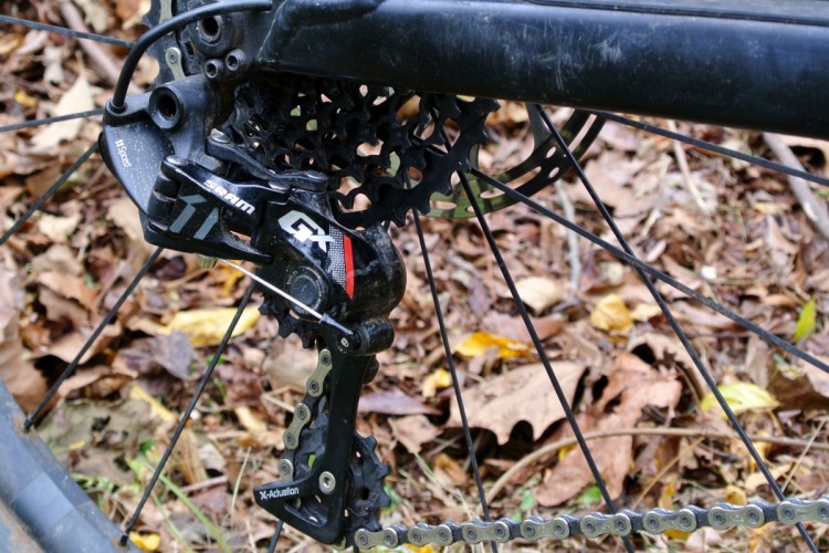 Close-up of a SRAM GX derailleur attached to a bicycle, with a visible chain and cassette set against a background of fallen autumn leaves.