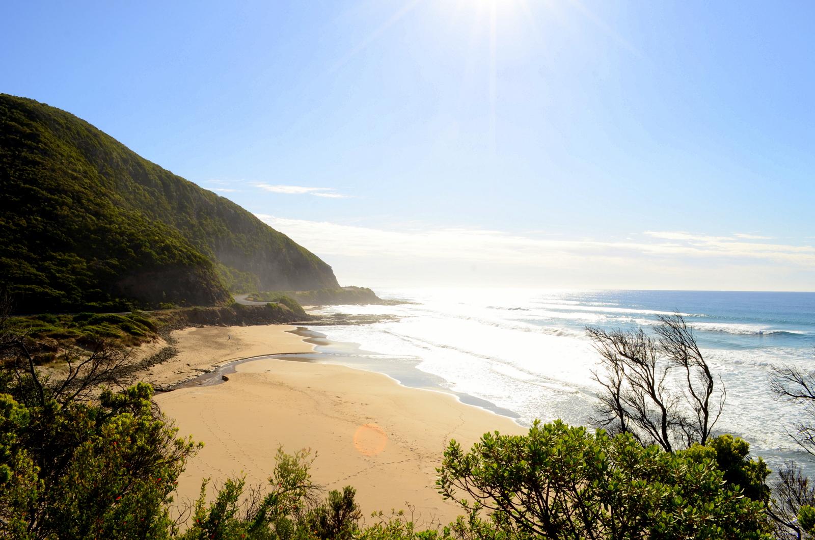 A scenic coastal view featuring a sandy beach bordered by a hill covered in lush greenery. The sunlight glimmers on the ocean waves, while gentle clouds scatter across the blue sky. A small stream can be seen flowing towards the beach, creating a serene and tranquil atmosphere. Forrest Mtb Trails mountain bike trail.