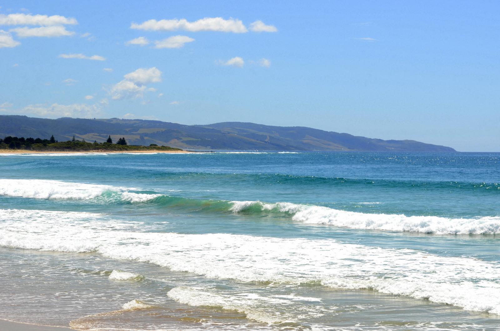 A serene coastal scene featuring gentle waves lapping at the sandy shore, with a backdrop of rolling hills and a clear blue sky dotted with a few clouds. The water is a vibrant blue, reflecting the sunlight, creating a serene and inviting atmosphere. Forrest Mtb Trails mountain bike trail.
