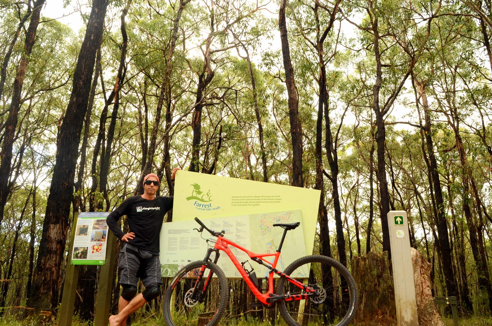 A person in a black shirt and sunglasses stands next to a mountain bike and a trail map sign in a forested area. The background features tall trees and a sign detailing mountain bike trails in the Forrest region. Forrest Mtb Trails mountain bike trail.