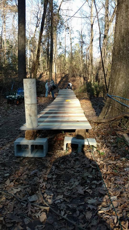 A partially constructed wooden path stretches through a wooded area, supported by concrete blocks and wooden beams. Two individuals can be seen working on the path, surrounded by fallen leaves and trees in a natural setting. Bringle Lake Mountain Bike Trail System mountain bike trail.