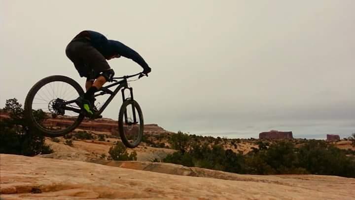 A mountain biker performing a jump on rocky terrain, with a scenic backdrop of cliffs and sparse vegetation under a cloudy sky. Navajo Rocks mountain bike trail.