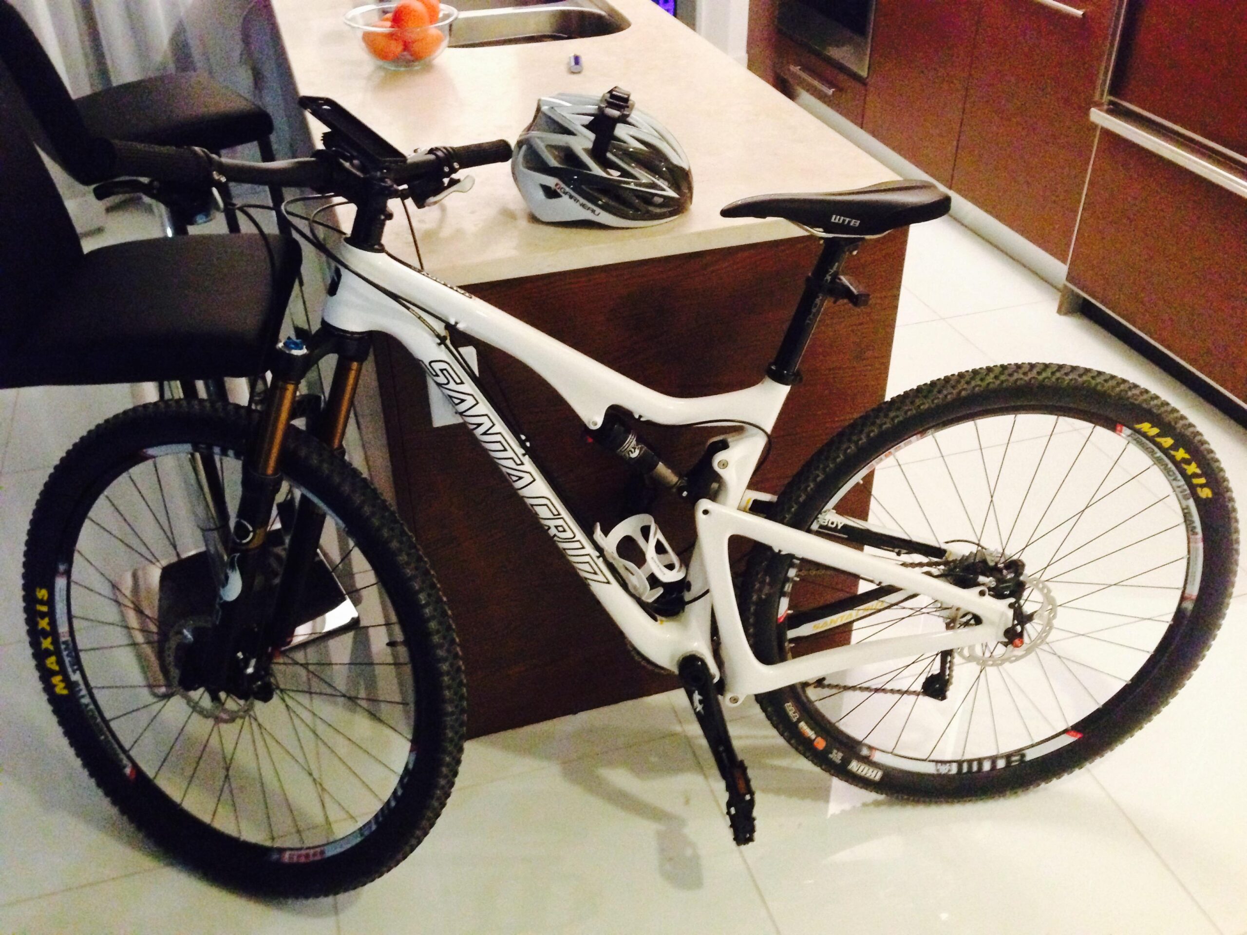 Santa Cruz Tallboy Carbon: A white mountain bike with a sleek design is parked next to a kitchen island. A black helmet rests on the countertop beside the bike. In the background, there are bar stools and a bowl of oranges, showcasing a modern kitchen setting. The bike features thick tires suitable for off-road riding.