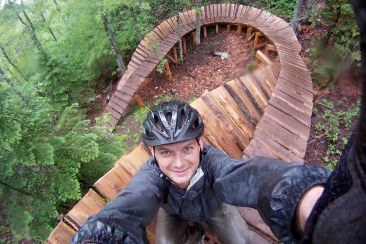 A person wearing a helmet takes a selfie while standing on a wooden mountain biking trail that curves through a lush, green forest, with rain visible on their jacket and the trail.