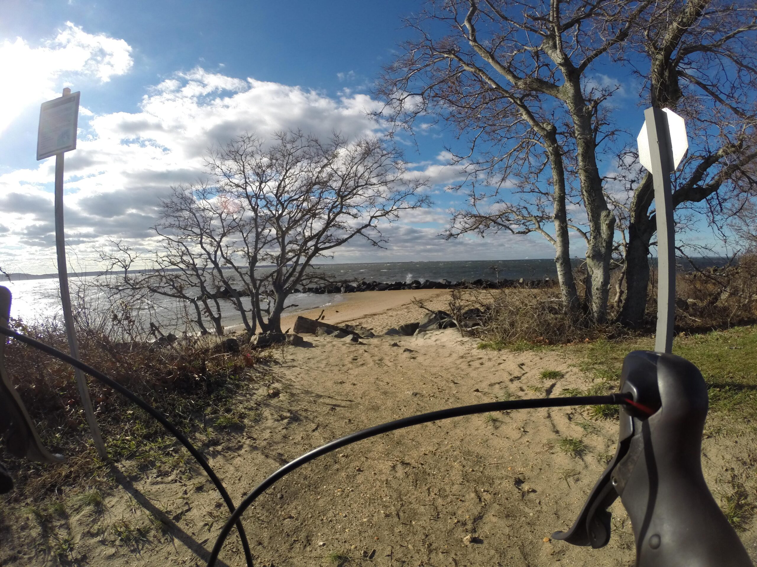 A scenic view of a coastline with sandy beach and water, framed by bare trees and a signpost. In the foreground, a bicycle handlebar is partially visible, suggesting a biking perspective. The sky is partly cloudy, with sunlight reflecting off the water. Sandy Hook Multi Use Path mountain bike trail.