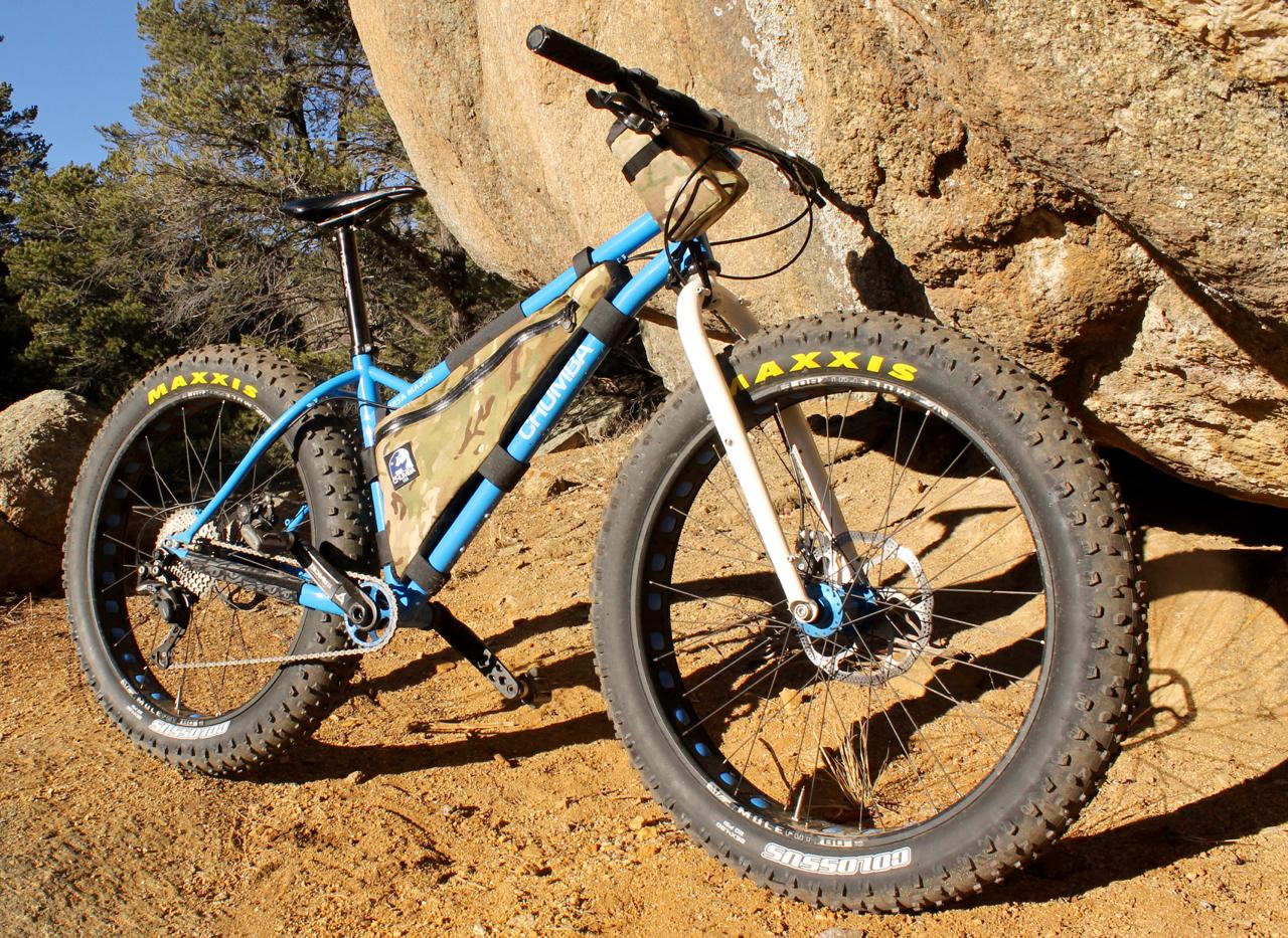 Chumba Ursa Major: A blue fat tire bike with a camouflage frame bag, parked on sandy ground near large boulders in a forested area. The bike features Maxxis tires, a front suspension fork, and a chain drive system. Sunlight illuminates the scene, showcasing the natural surroundings.