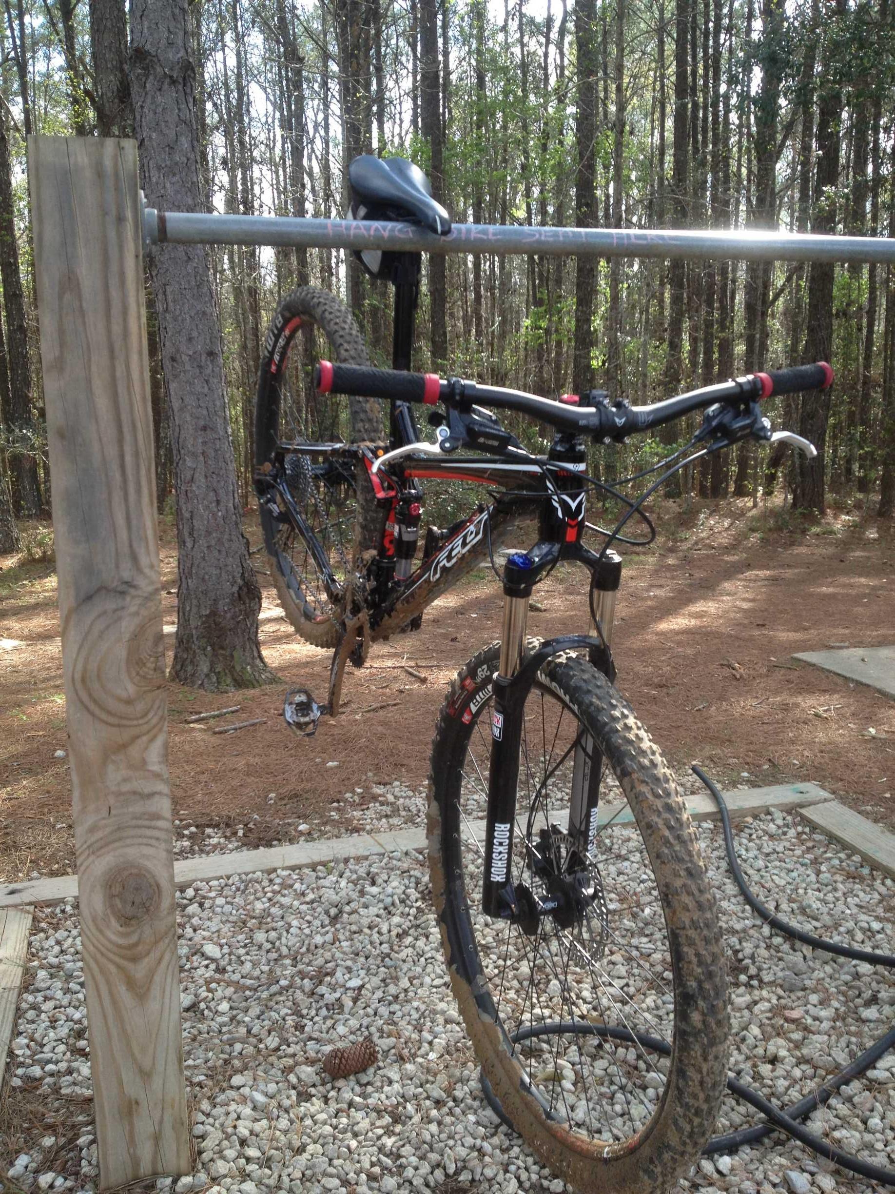 A mountain bike suspended on a metal rack in a wooded area. The bike has a dirty frame and tires, indicating recent use on a trail. The background features tall trees and a mix of soil and gravel underfoot. A wooden post is visible next to the bike. Redbug mountain bike trail.