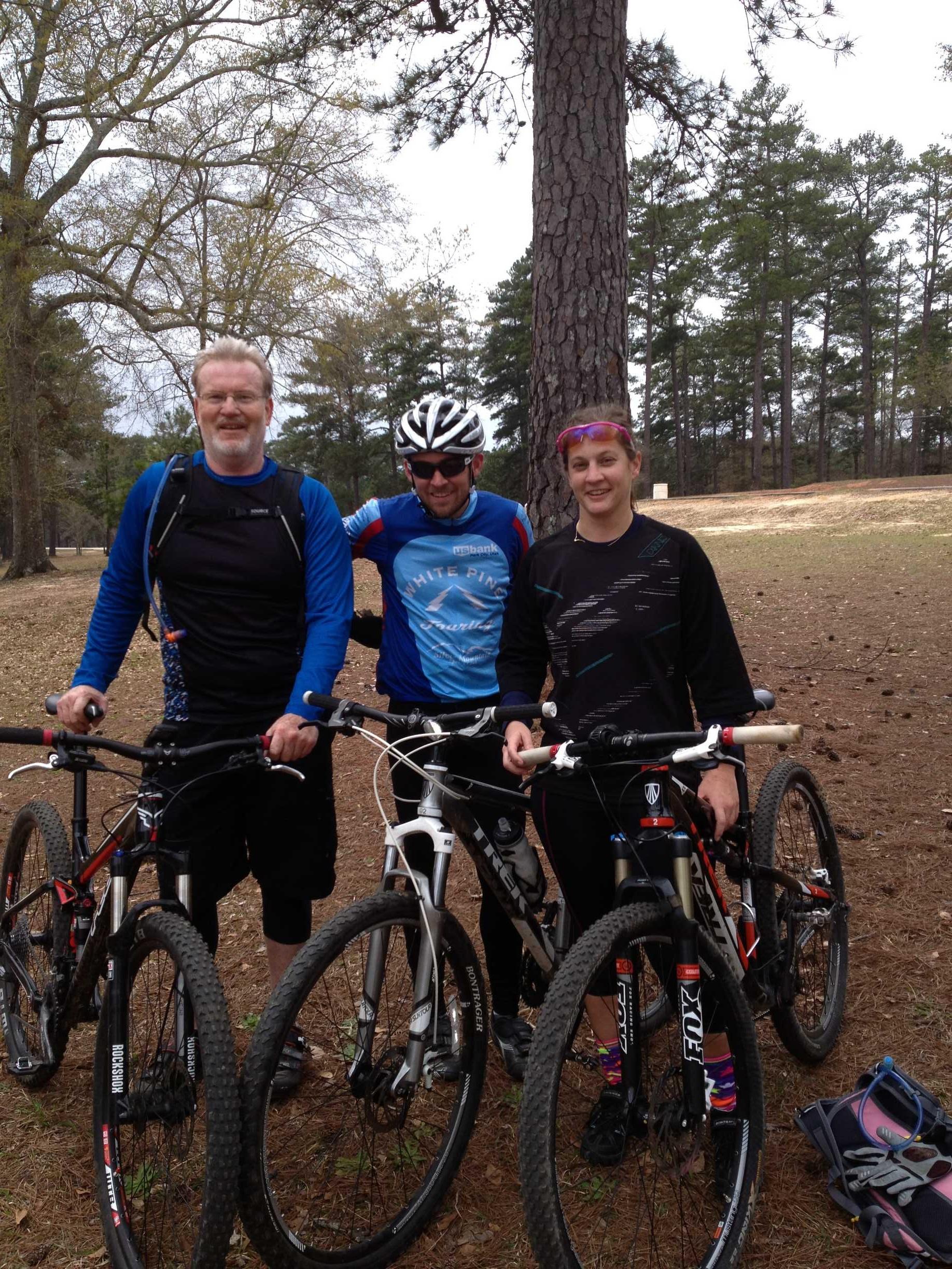 Three mountain bikers pose together outdoors, each holding their bicycles. The group stands in a wooded area, with tall trees in the background. The first person, on the left, has gray hair and wears a black and blue biking outfit. The second person, in the middle, wears a blue jersey and a helmet, while the third person, on the right, has short hair and wears a black outfit with colorful accents. All three appear to be enjoying their time in nature. Flat Rock Park mountain bike trail.