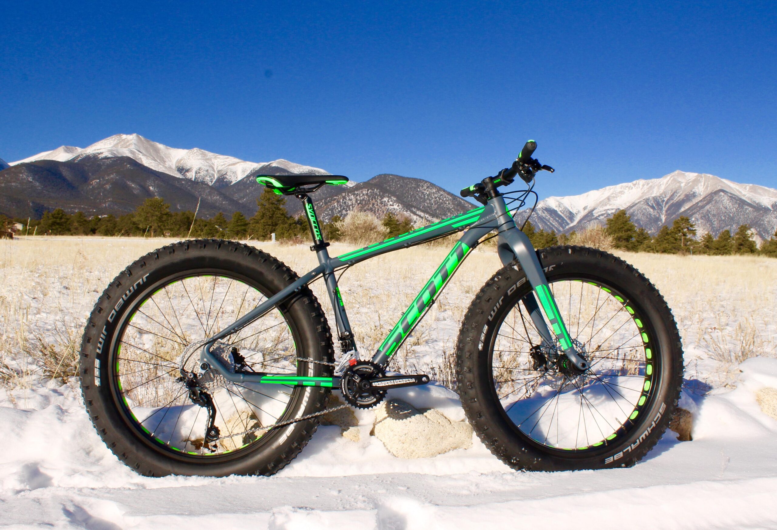 Scott Big Jon: A green and gray fat bike is parked on snowy ground, with a backdrop of snow-capped mountains and a clear blue sky. The bike features wide tires designed for snow and rugged terrain.