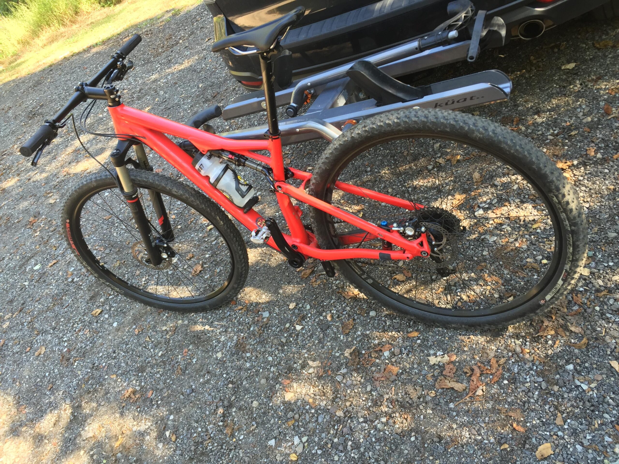 Specialized Epic Comp 29er: Close-up of a red mountain bike parked on a gravel surface, equipped with a water bottle holder and featuring knobby tires suitable for off-road cycling. The background shows greenery and part of a vehicle.