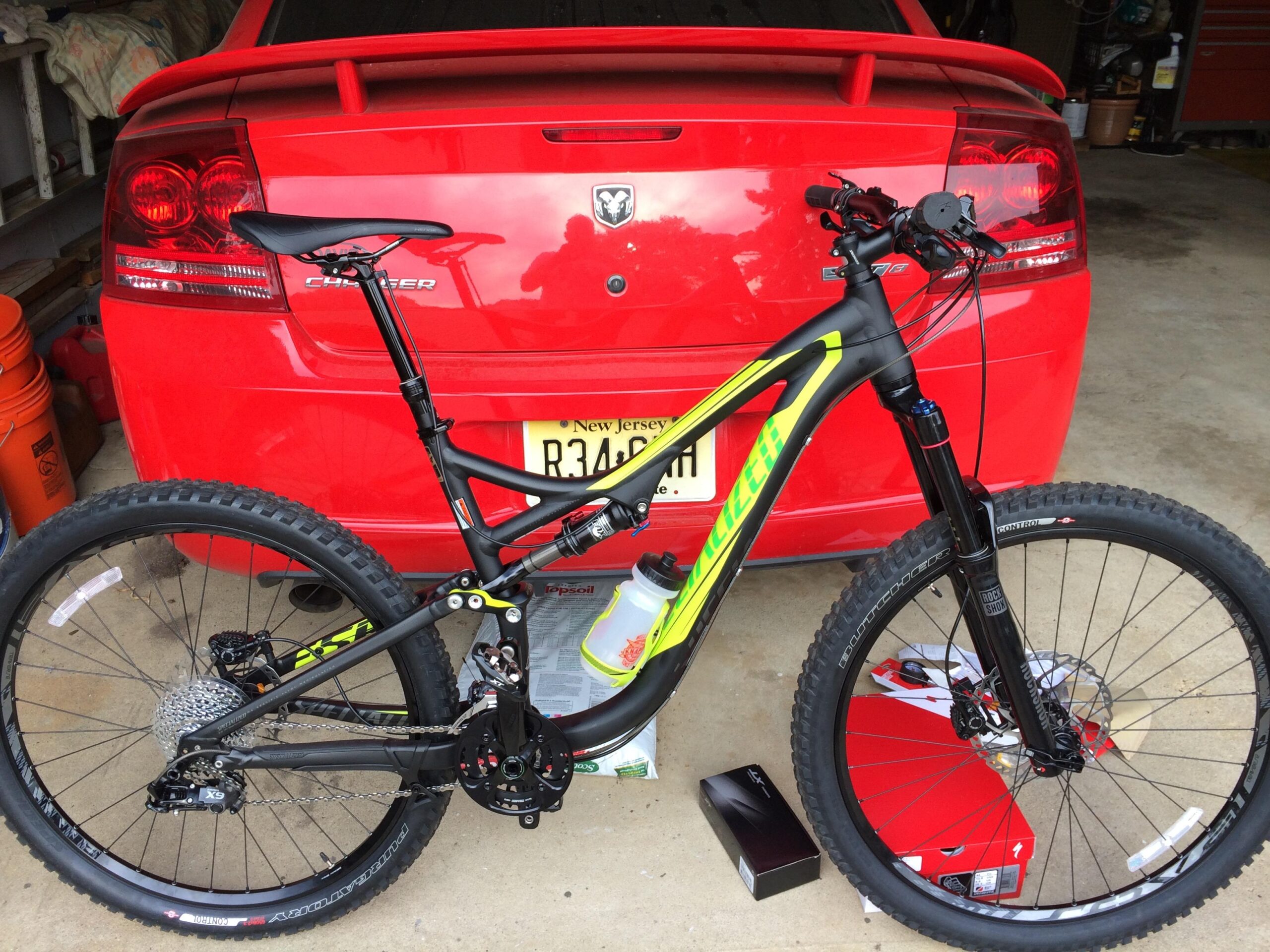 Specialized FSR: A black and yellow mountain bike is positioned in front of a red Dodge Charger in a garage setting. The bike features thick tires and a modern design, with components visible such as the suspension and gear system. Behind the bike, various items and containers are slightly obscured, while the focus remains on the bike and the car.