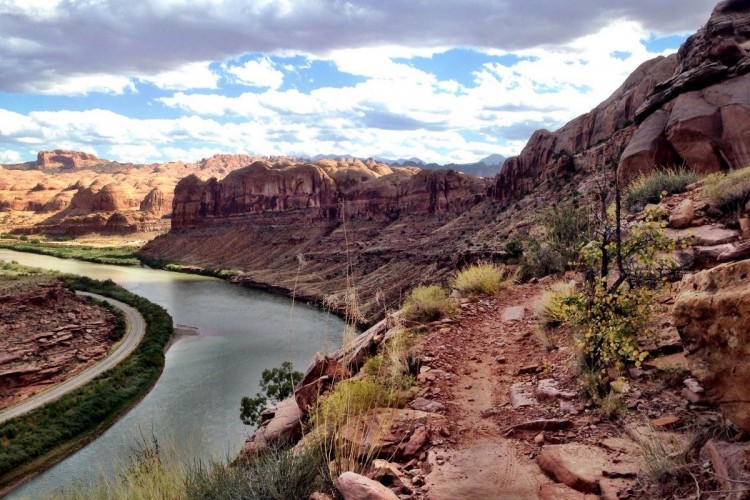 Scenic view of a winding river bordered by rocky cliffs and rolling hills under a partly cloudy sky. The foreground features a dirt path surrounded by dry vegetation and shrubs, leading to the edge of the landscape.