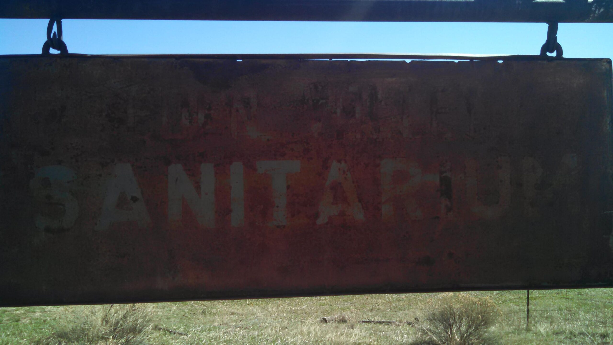 Old, rusted sign partially obscured, with the word "SANITARIUM" visible, hanging against a clear blue sky. The background features grassy terrain with low shrubbery. Eden Valley Spur mountain bike trail.