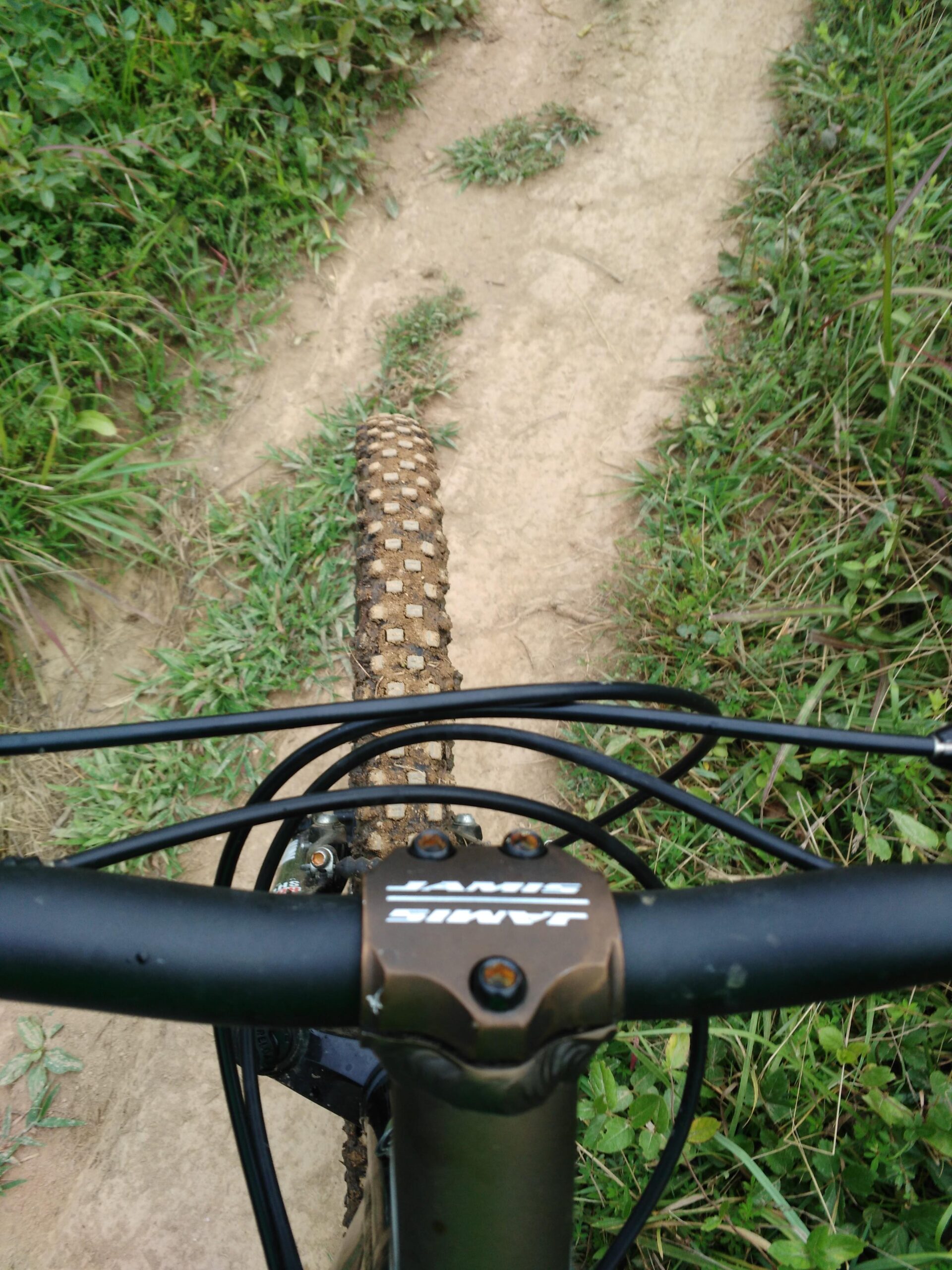 Trek 4500: Aerial view of a mountain bike's handlebars and front tire on a dirt trail, surrounded by green grass and foliage. The ground shows a sandy path with some vegetation.