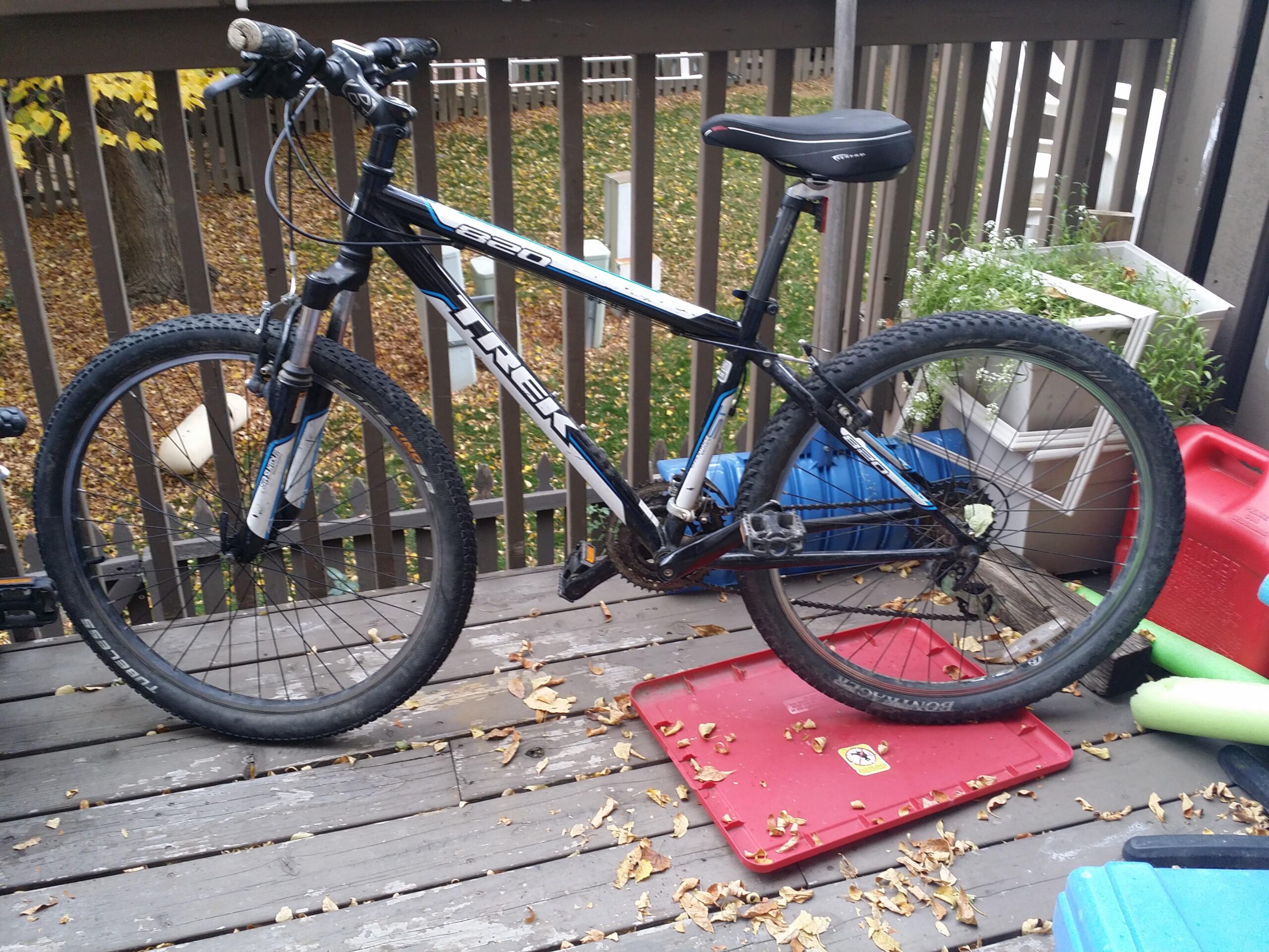 Trek 820: A black Trek mountain bike is parked on a wooden deck, surrounded by fallen leaves. The bike features a front suspension fork and knobby tires. Nearby, there are colorful plastic storage containers and a planter with greenery. The background shows a fenced area with some grass partially covered by leaves.