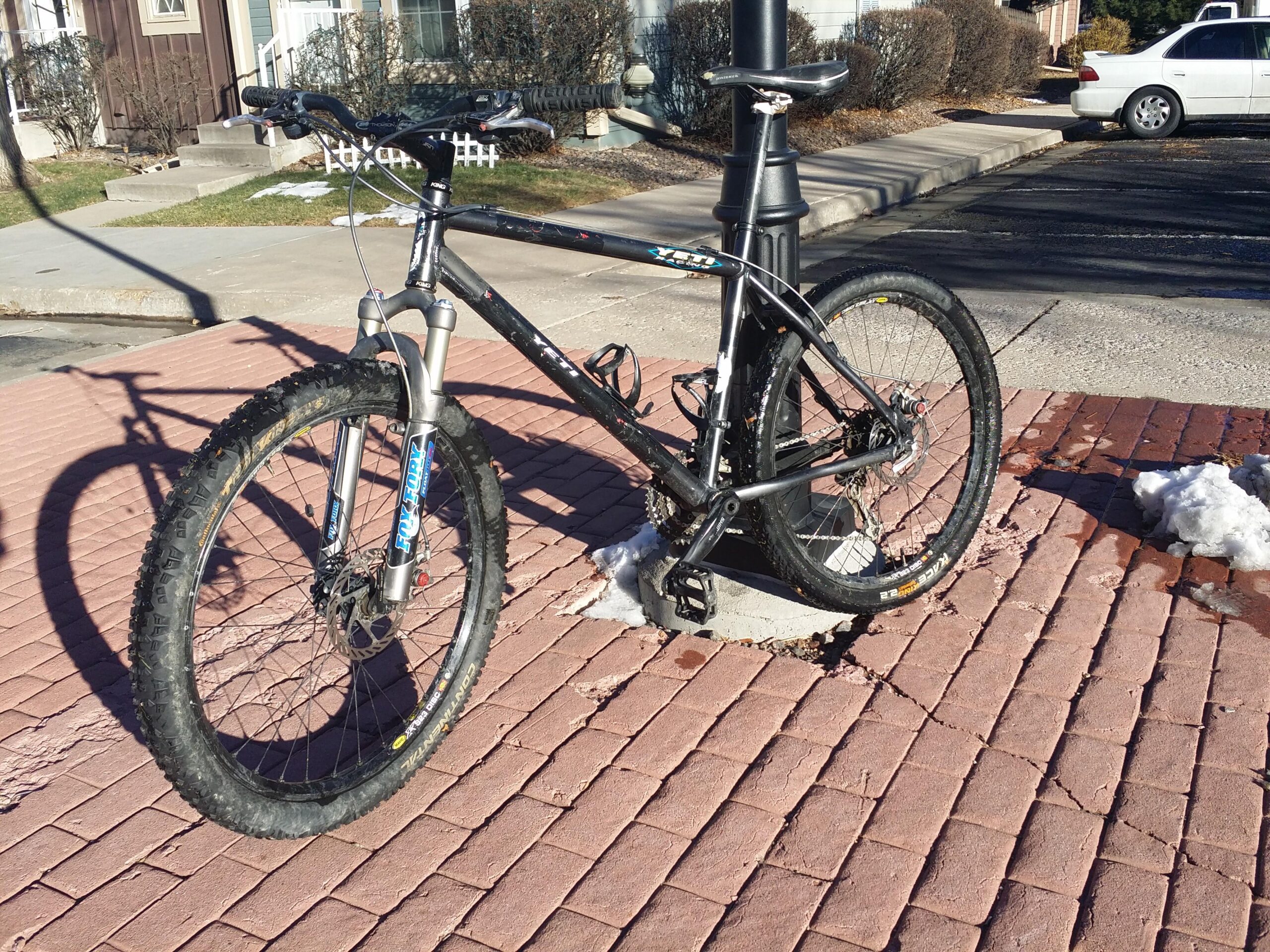 Kona Caldera: A black mountain bike is parked against a lamppost on a brick pavement. The bike features thick tires, a sturdy frame, and visible components such as brakes and pedals. In the background, there are patches of snow and a white car parked on the street. The setting appears to be in a residential area.