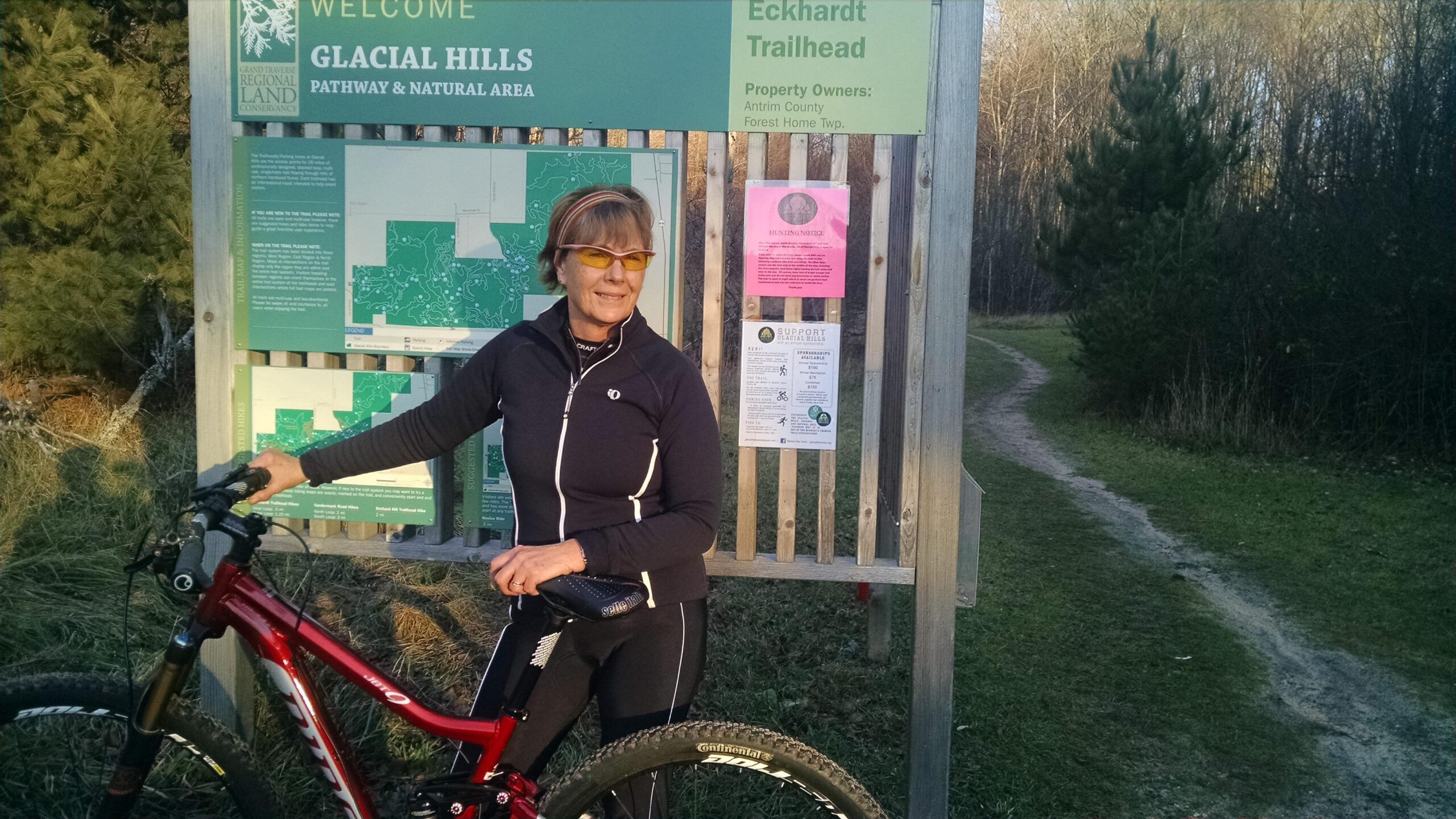A woman stands next to a trailhead sign at the Glacial Hills Pathway and Natural Area, holding her mountain bike. She is wearing sporty attire and sunglasses, and the background features trees and a trail leading into the woods. The sign includes maps and information about the area. Arcadia Dunes mountain bike trail.
