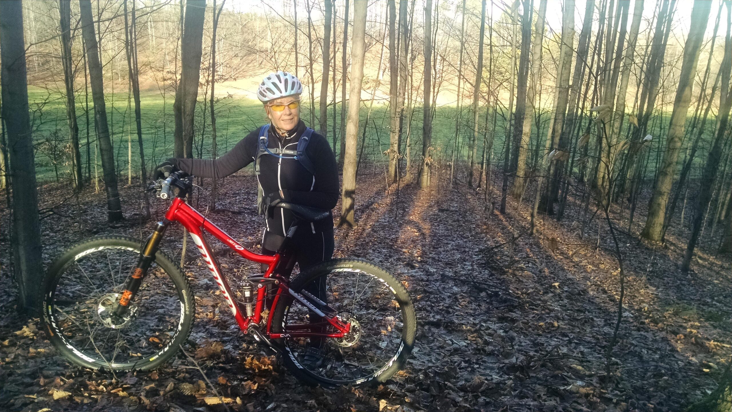 A cyclist stands beside a red mountain bike in a forested area. The rider is wearing a helmet and sunglasses, dressed in a black cycling outfit, with trees and fallen leaves in the background. Sunlight filters through the trees, illuminating the scene with a warm glow. Arcadia Dunes mountain bike trail.