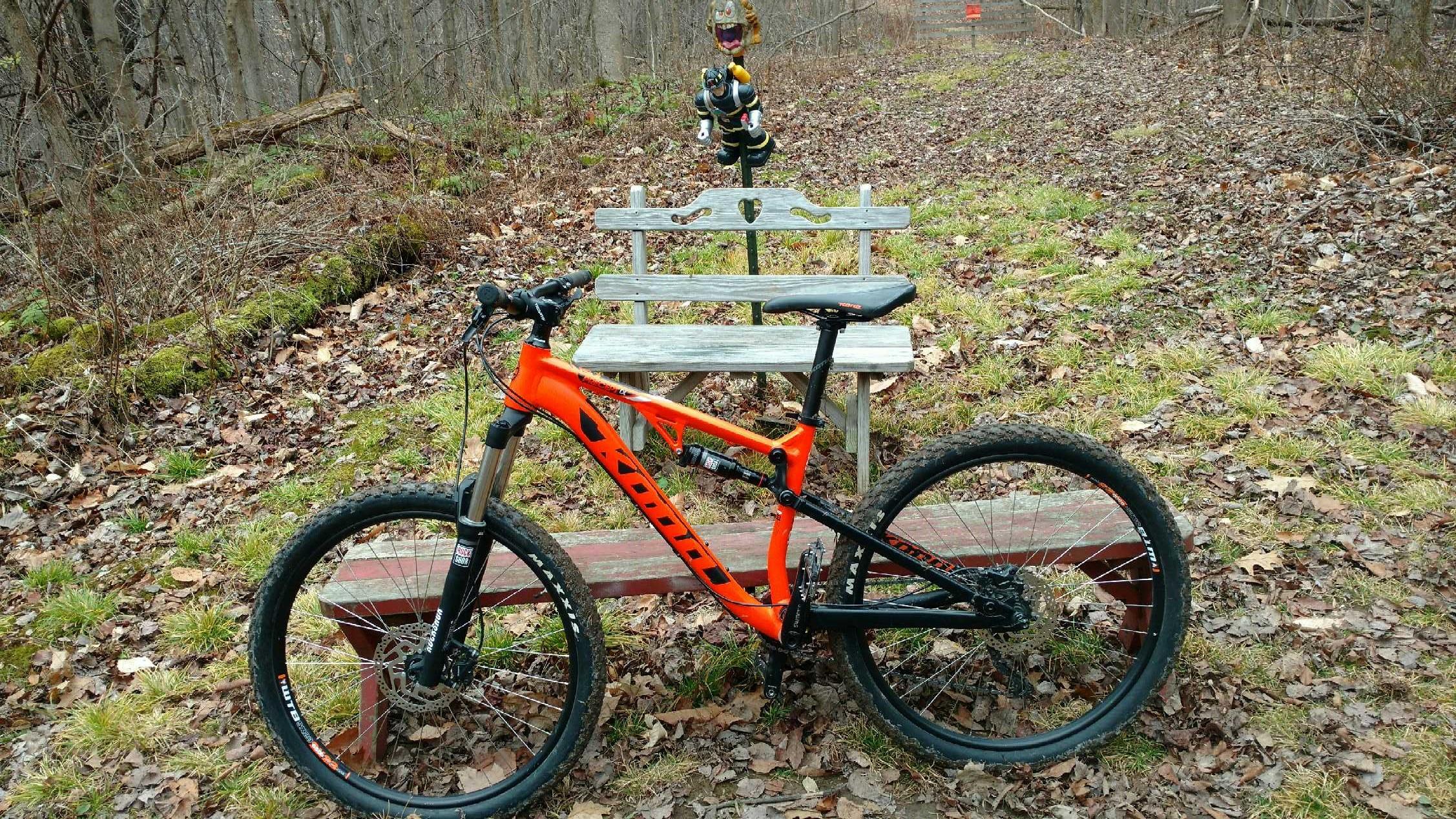 A vibrant orange mountain bike is positioned on a leaf-covered trail, with a wooden park bench in the background. A playful figure resembling a cartoon character stands on a pole behind the bench, adding a whimsical touch to the natural setting. Trees are visible in the background, indicating a wooded area. Great Bear mountain bike trail.