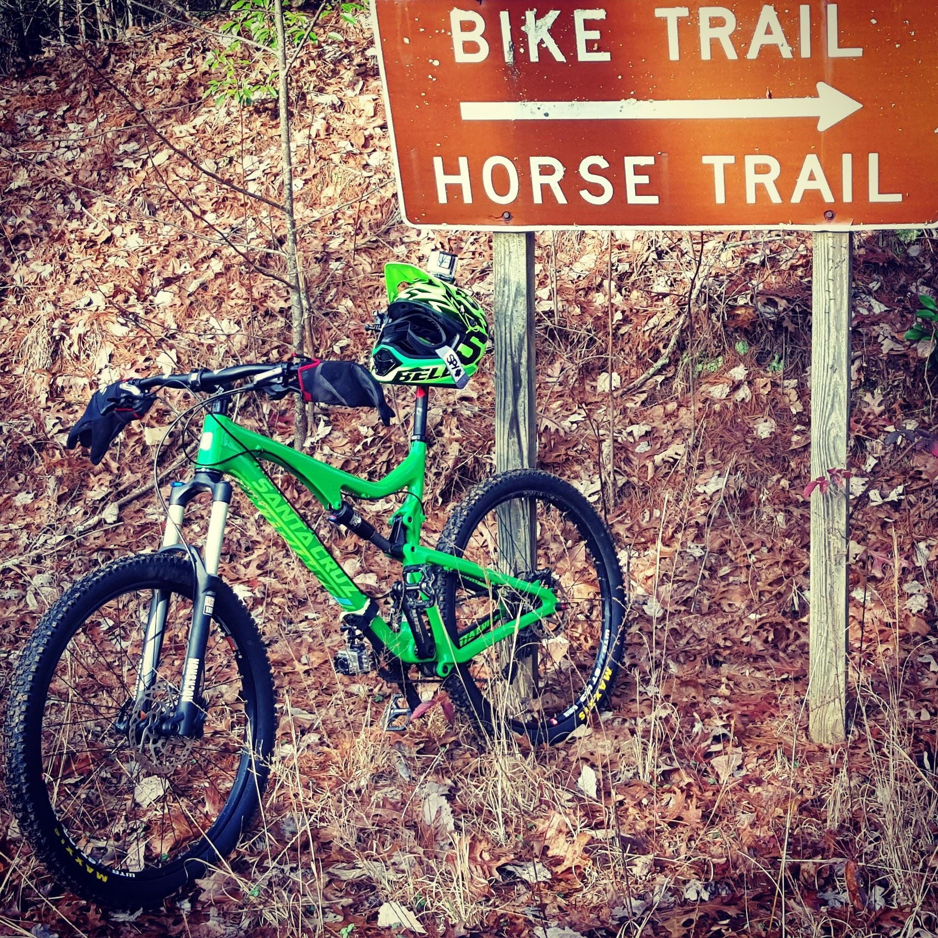 A bright green mountain bike with a helmet resting on it, positioned next to a sign indicating directions for a bike trail and a horse trail, surrounded by fallen leaves and trees. Bull / Jake Mountain mountain bike trail.