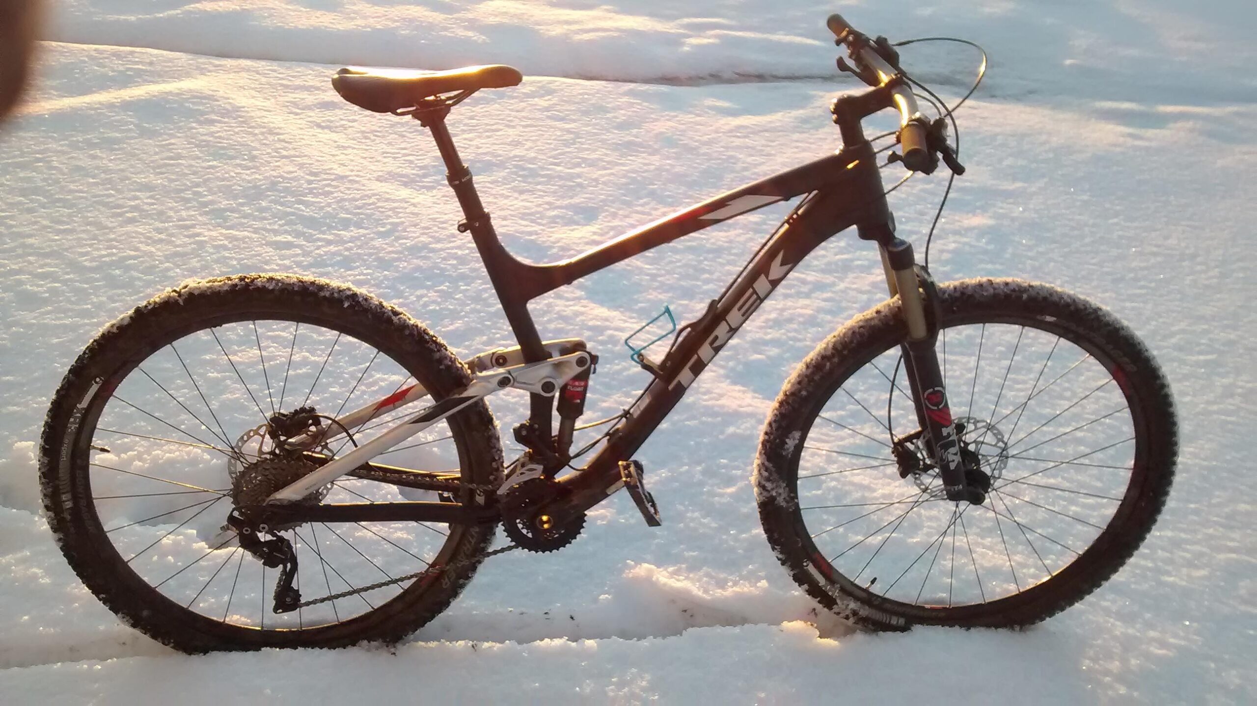 Trek Fuel EX 8 29: A mountain bike resting on a snowy surface, featuring a black and silver frame with thick, knobby tires. The sunlight casts a warm glow on the bike, highlighting its details, while the surrounding snow reflects a sparkling texture.