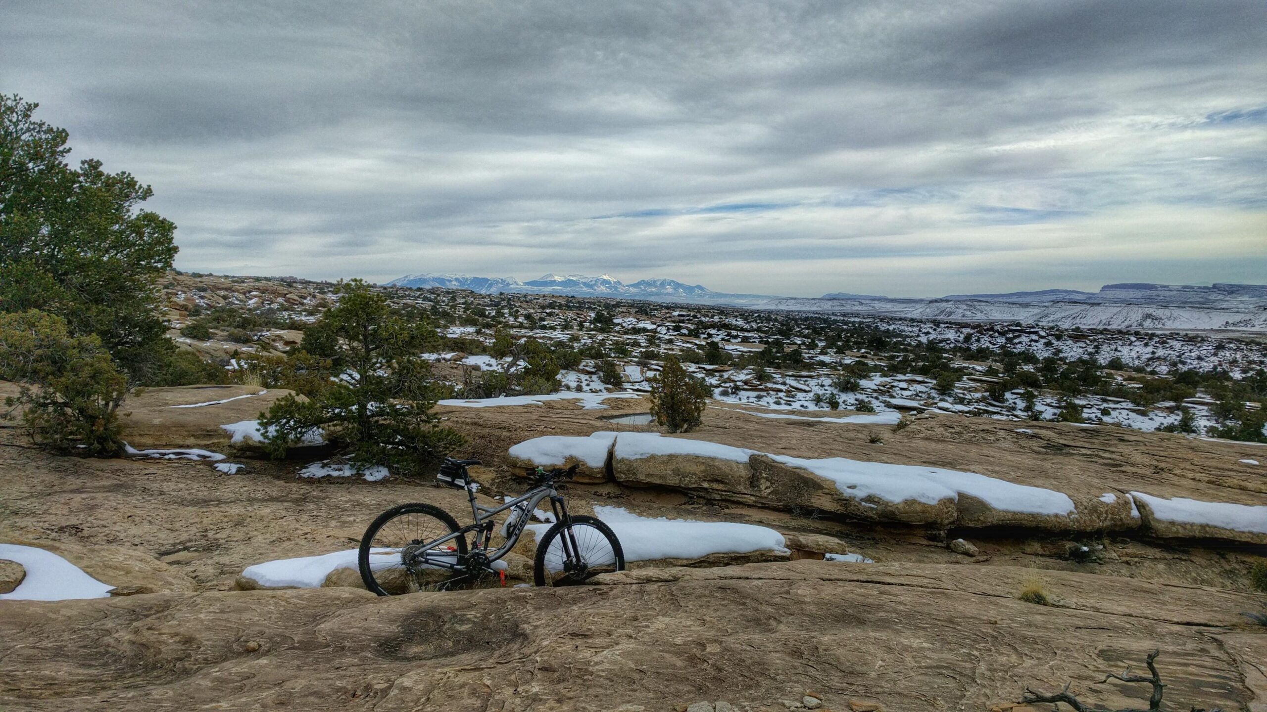 A mountain bike resting on rocky terrain, surrounded by sparse vegetation and patches of snow, with a cloudy sky overhead and distant mountains in the background. Klondike Bluffs mountain bike trail.