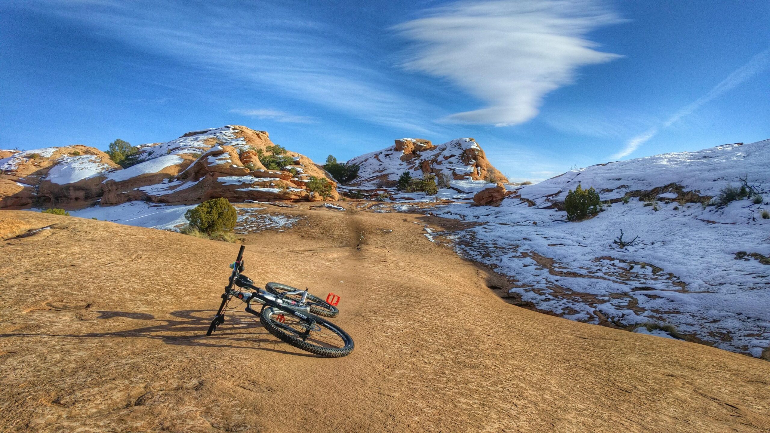 A mountain bike resting on a rocky, snow-dusted trail surrounded by red rock formations and blue skies. Slickrock mountain bike trail.