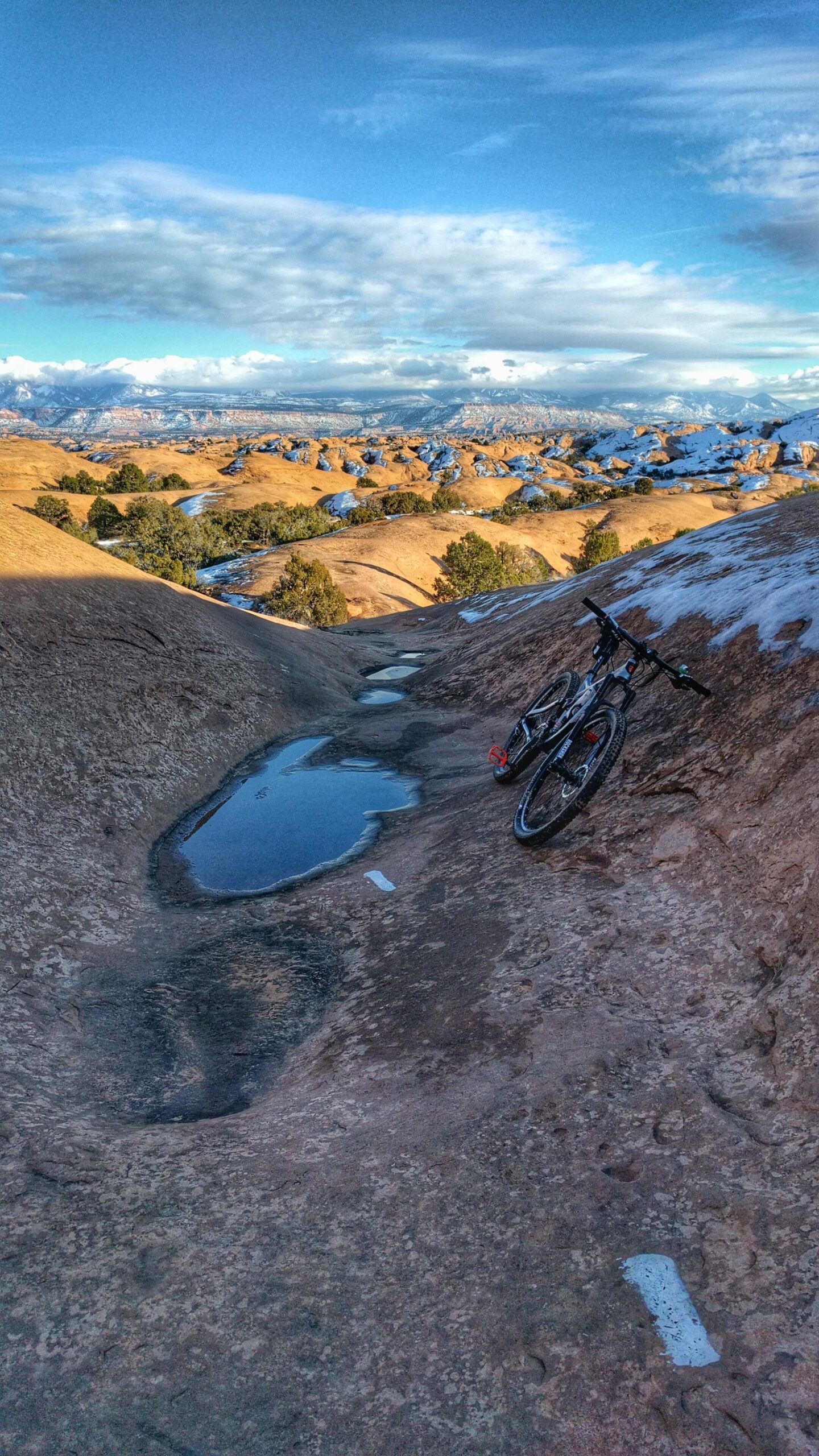 A mountain bike leaning against a rocky terrain with two puddles reflecting the sky. In the background, rolling hills and mountains can be seen under a partly cloudy blue sky. The landscape features a mix of rocky formations and patches of snow. Slickrock mountain bike trail.