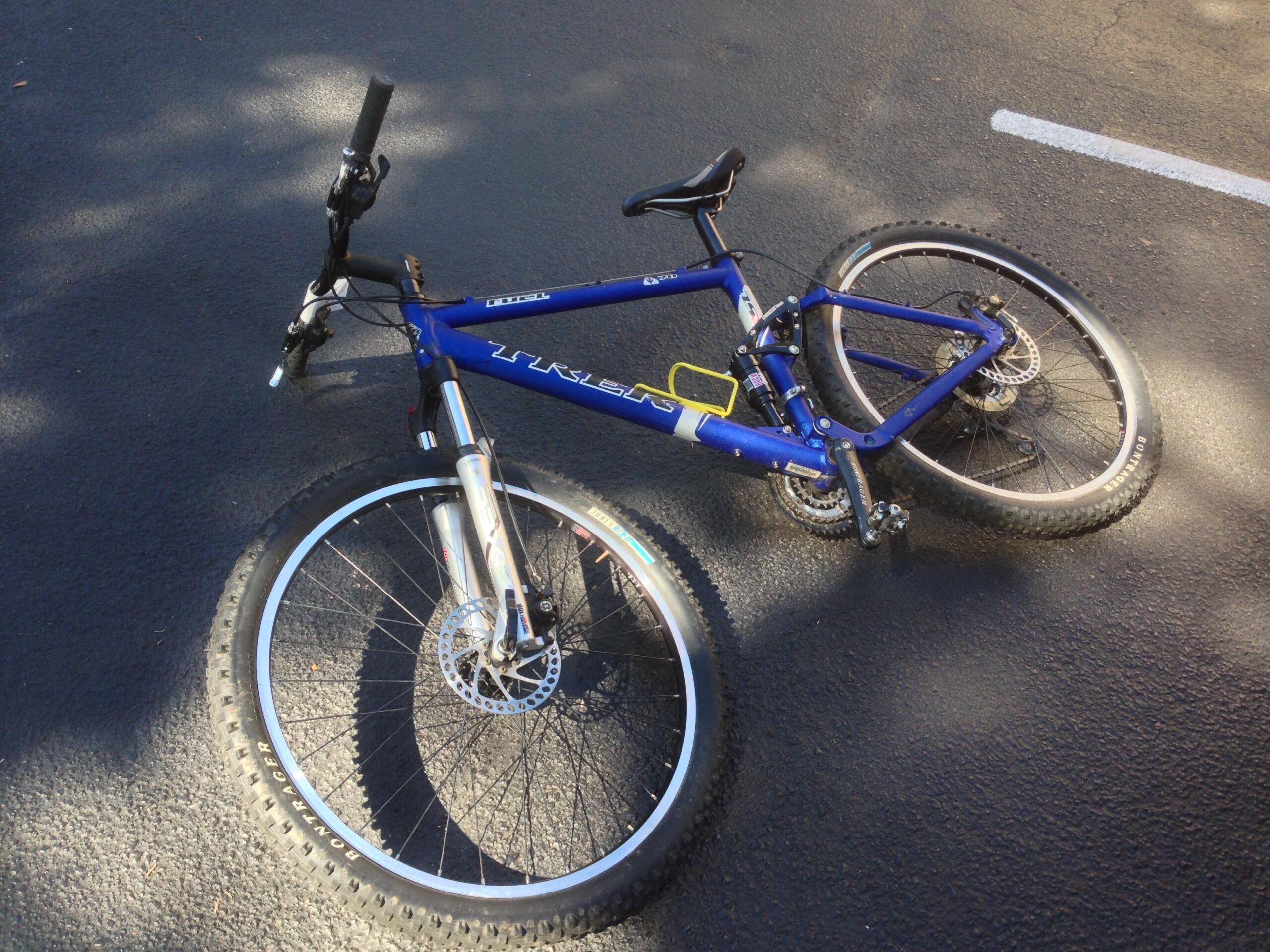 Trek Fuel EX: A blue mountain bike lying on a black asphalt surface, with one wheel raised off the ground. The bike features a prominent front suspension fork, disc brakes, and a geometric frame design typical of mountain bikes. Sunlight casts a shadow on the ground, hinting at a warm day.