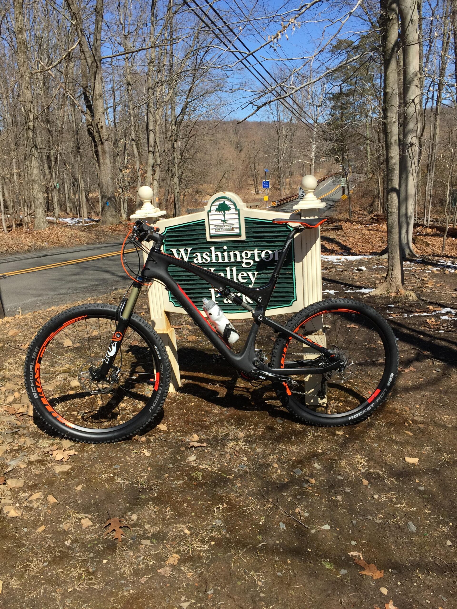 Scott Genius 710: A mountain bike parked beside a sign for Washington Valley, surrounded by bare trees and gravel. The scene is set on a sunny day with clear blue skies.