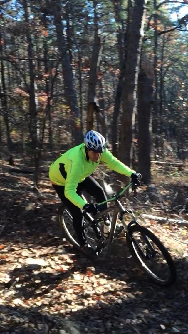 A person wearing a bright yellow jacket and a helmet is riding a mountain bike on a forest trail covered with leaves. The rider is leaning forward, navigating a turn among the trees in a wooded area. The sunlight filters through the branches, creating dappled light on the ground. Iron Mountain mountain bike trail.