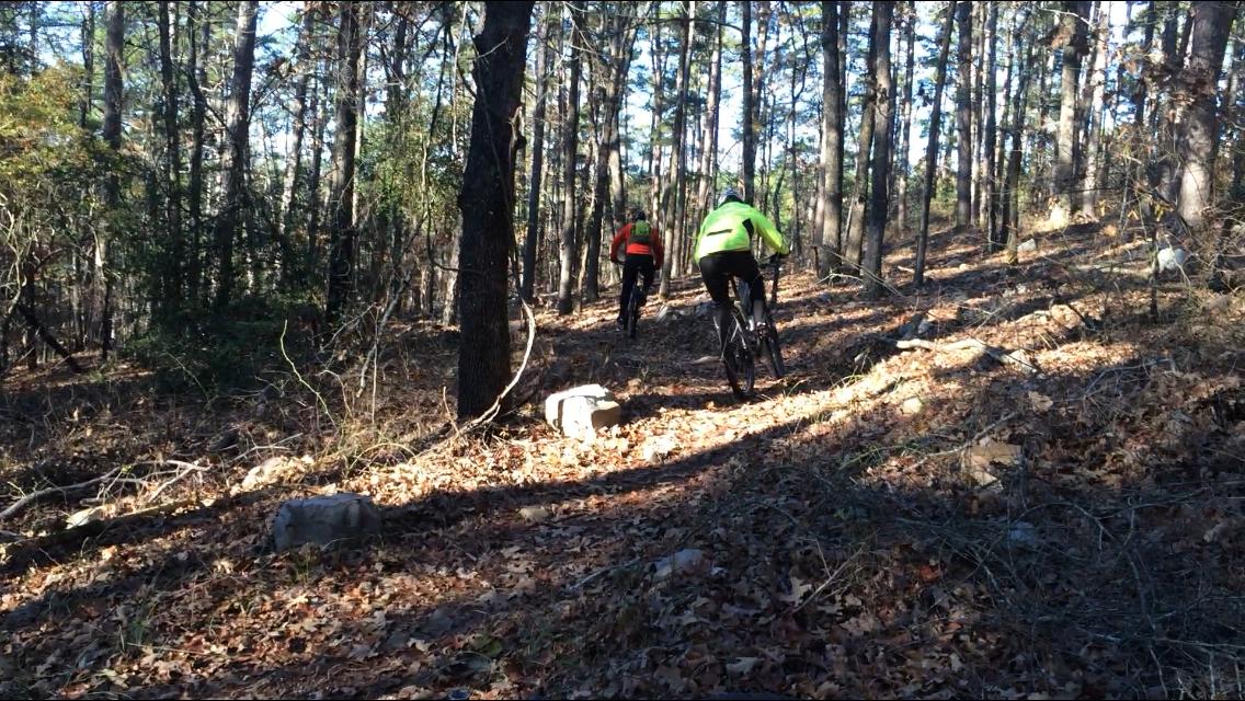 Two mountain bikers navigate a wooded trail covered with leaves and rocks. The riders, dressed in bright jackets, are biking uphill in a forested area with tall trees and dappled sunlight filtering through the canopy. Iron Mountain mountain bike trail.