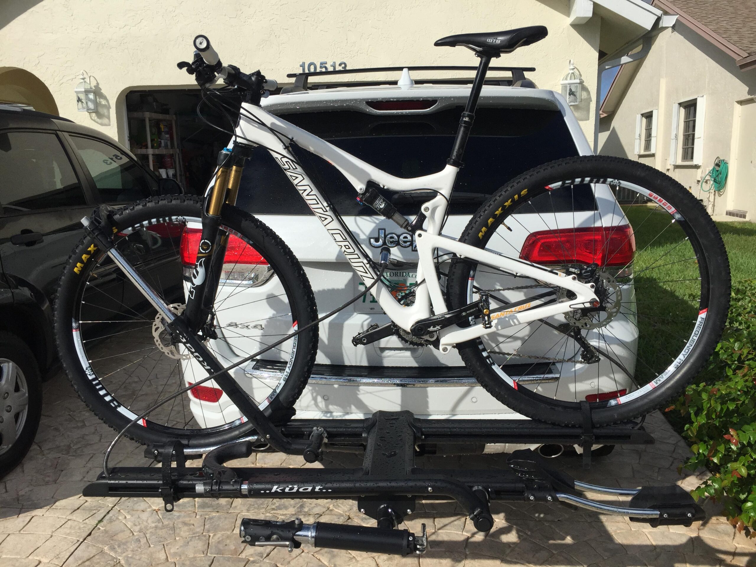 Santa Cruz Tallboy Carbon: A mountain bike mounted on a rear bike rack attached to a white SUV, with a garage in the background. The bike features a sleek design with a white frame and prominent brand markings, and is secured with a locking mechanism on the rack.