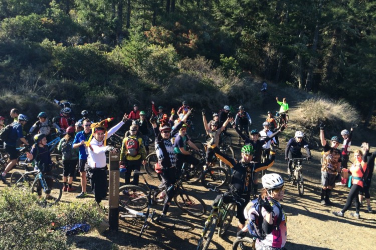 A large group of mountain bikers, both adults and children, gather on a dirt trail surrounded by trees. Many riders are wearing helmets and colorful cycling gear. Some are waving and smiling at the camera, while others are chatting and enjoying the outdoor atmosphere. A signpost is visible in the foreground, pointing towards a nearby trail. The sun shines brightly, creating a vibrant and energetic scene.
