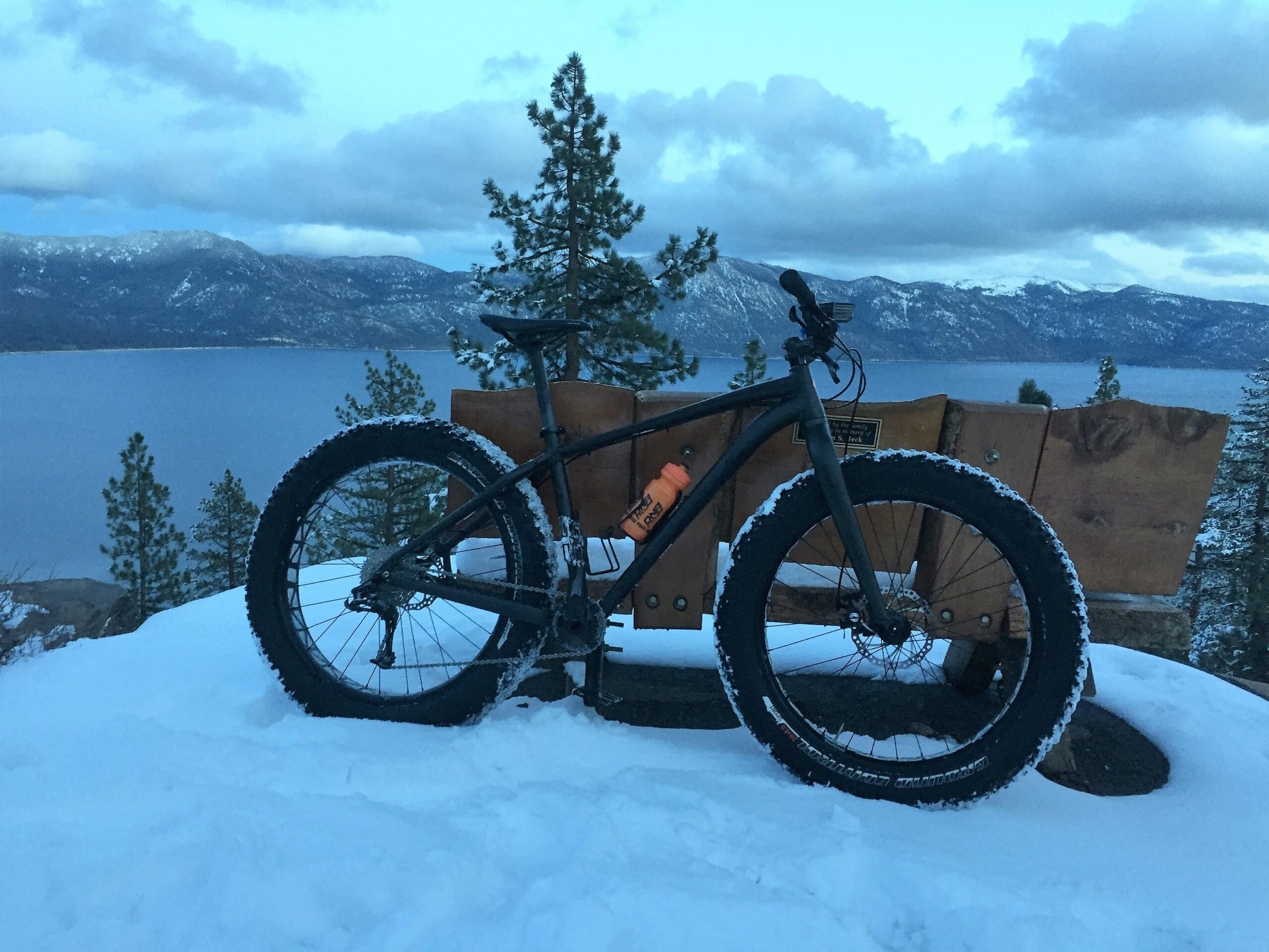 Specialized Fatboy: A fat tire bike parked on a snowy surface, with a wooden bench in the background overlooking a lake and mountains. The sky is cloudy and the light is soft, suggesting early morning or late afternoon. Pine trees are scattered around the scene.