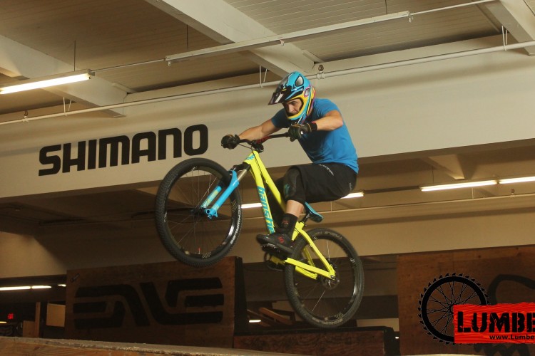A mountain biker in a blue shirt and helmet performs a jump in a bike park, with the Shimano logo visible in the background. The rider's bike is predominantly yellow and blue, showcasing the dynamic movement as the front wheel rises off the ramp.