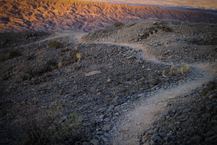 A winding dirt path meanders through a rocky landscape, surrounded by sparse vegetation. The mountains in the background are bathed in warm, golden light, highlighting their texture and contours against a clear sky.