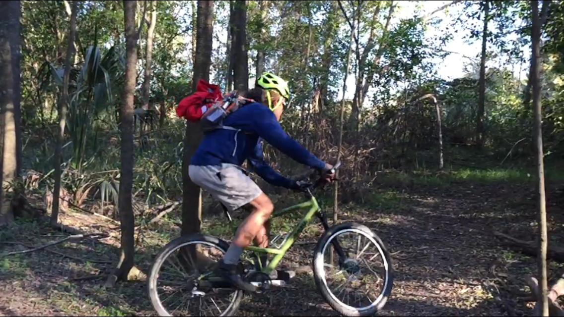 A person riding a green mountain bike on a dirt trail in a wooded area. They are wearing a yellow helmet and a blue long-sleeve shirt, with gray shorts. A red backpack is attached to their bike, and the scene features trees and underbrush in the background.