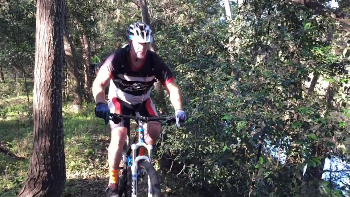 A mountain biker navigating a wooded trail, wearing a helmet and cycling gear, with trees and greenery surrounding the path.