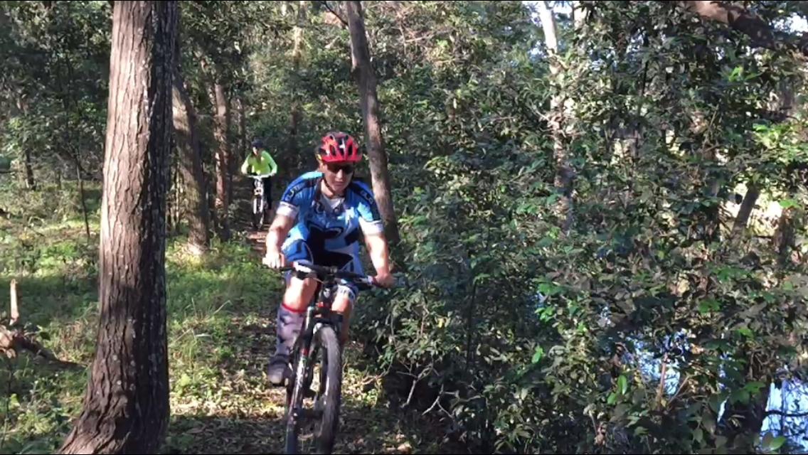 A mountain biker riding along a narrow trail through a dense, green forest. The biker is wearing a blue jersey, red helmet, and sunglasses, and appears focused on navigating the path. Another cyclist in a bright yellow jacket is seen in the background.